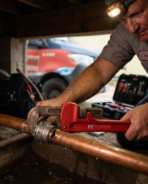 Mechanic using a red pipe wrench to tighten a copper pipe under a workbench
