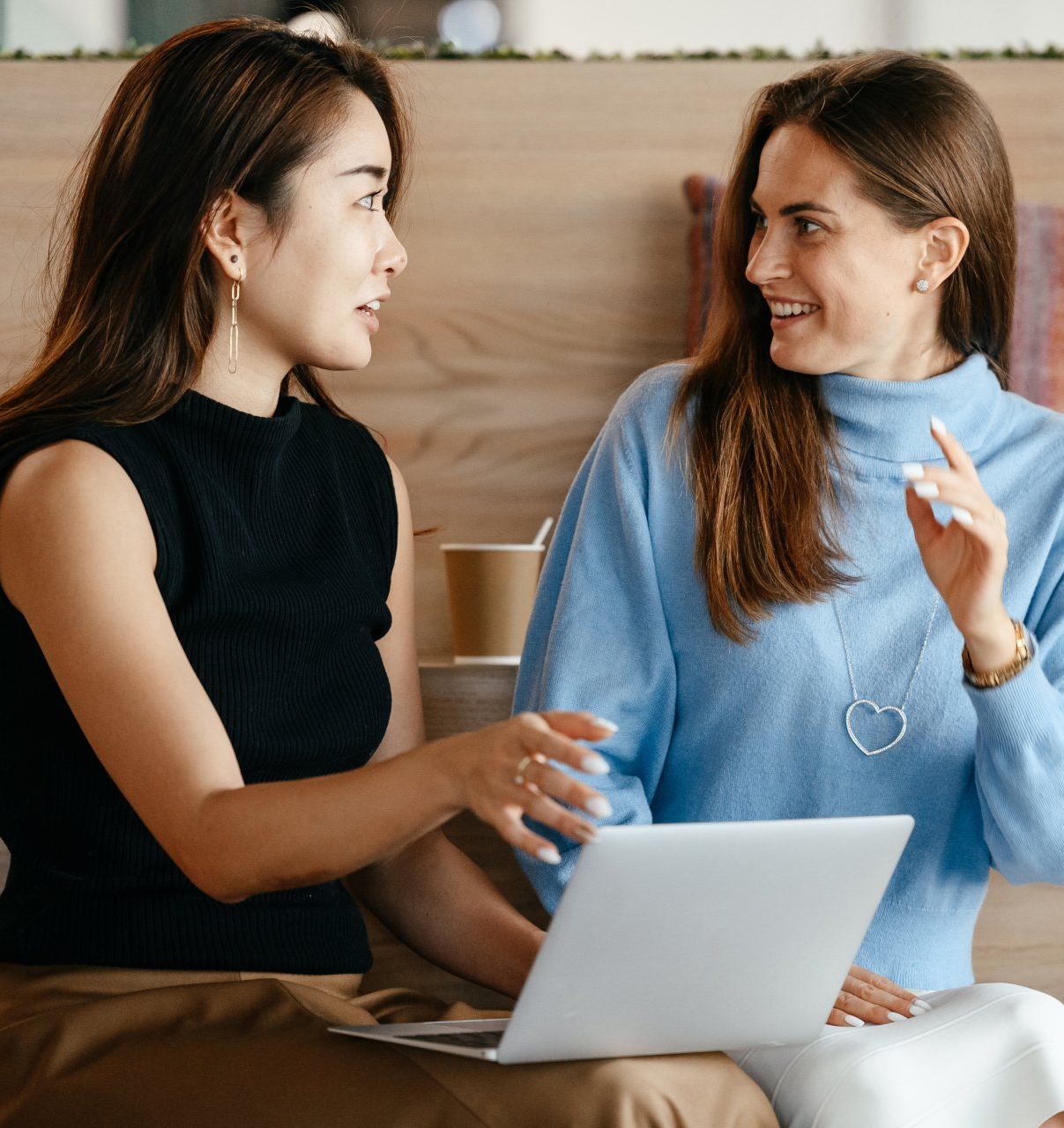 Two women are sitting at a table with a laptop and talking to each other