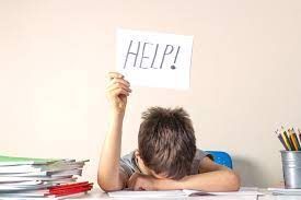 A young boy is sitting at a desk holding a sign that says `` help ''. Executive Functioning Coaching, ADHD