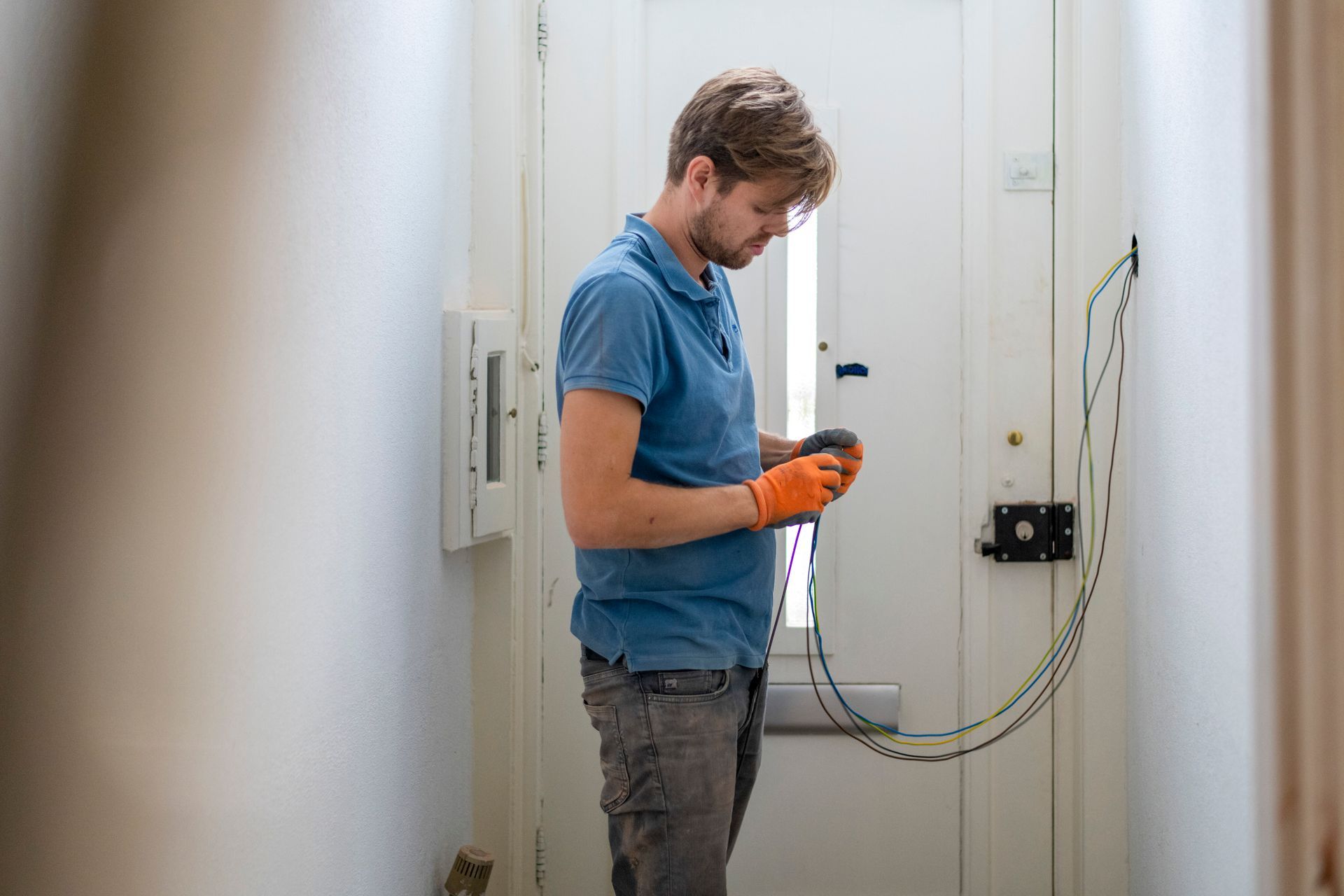 Man in blue shirt and gloves inspecting wires by a white door.