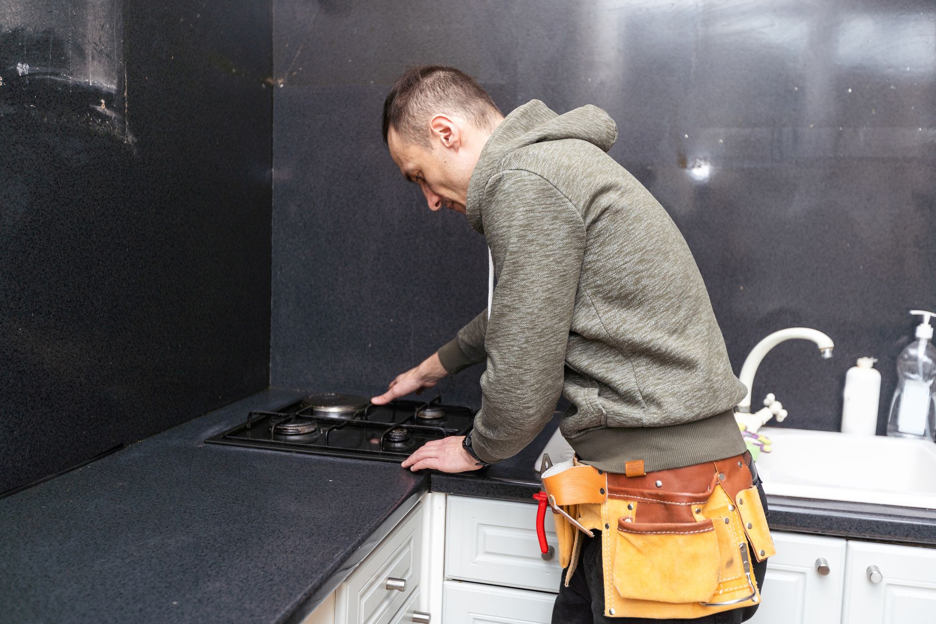Man in hoodie installing a gas cooktop in a black kitchen with white cabinets.