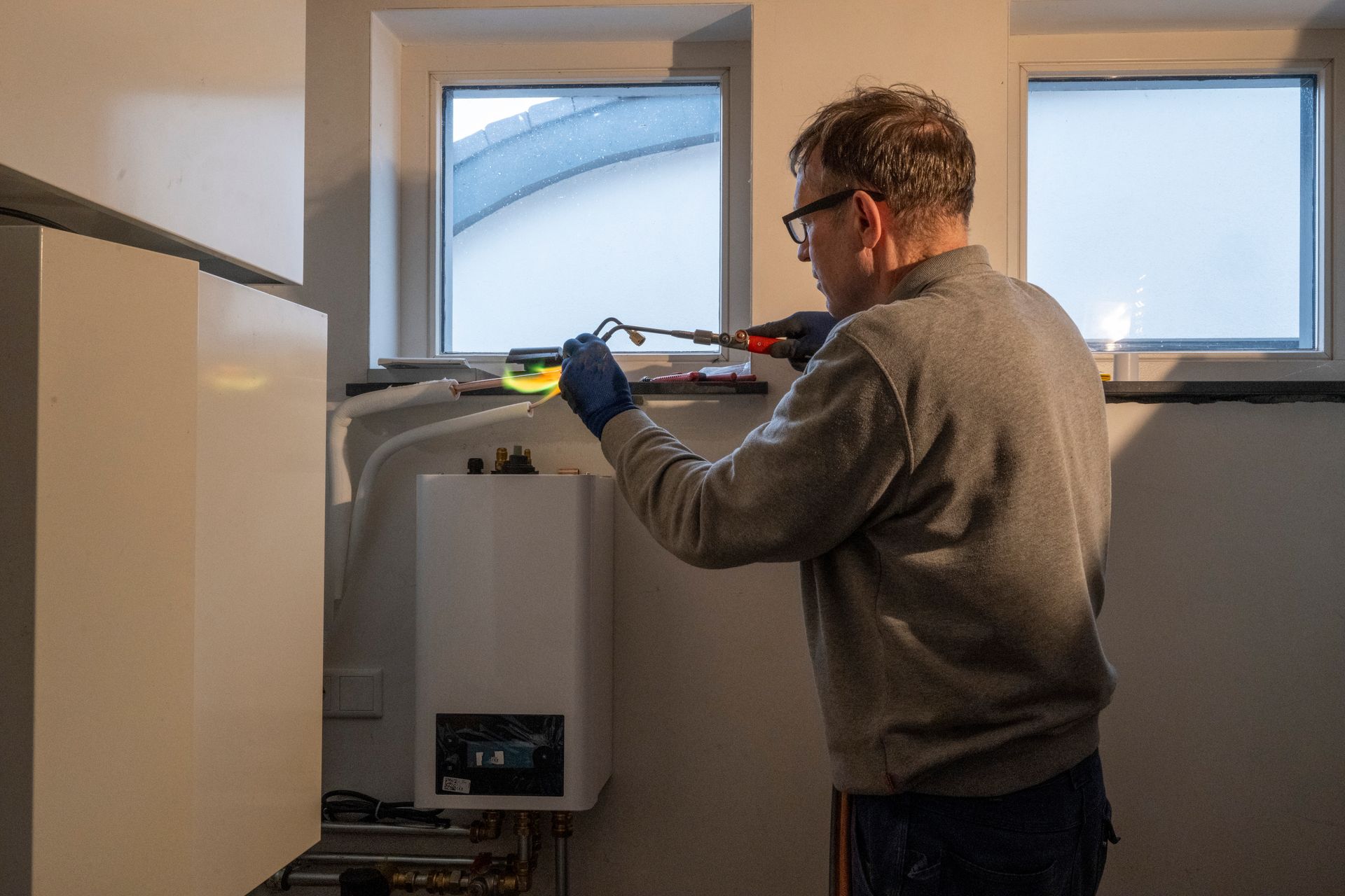 Man in gray sweater repairs a white appliance by a window, using a screwdriver.
