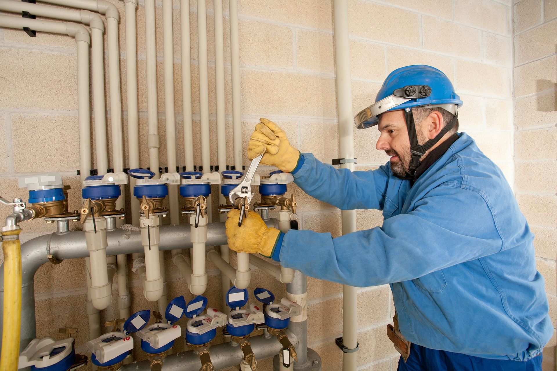 Plumber in blue jacket and hard hat working on pipes with a wrench.
