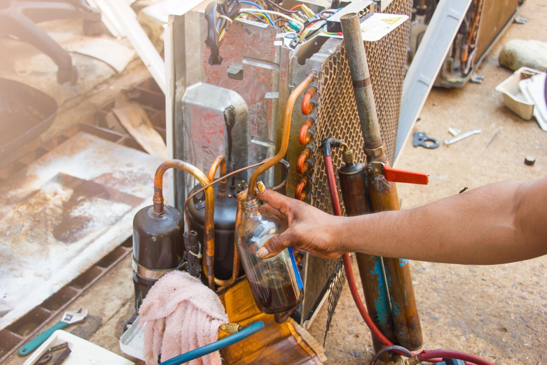 Person's hand pointing at AC unit components, likely during repair. Copper coils, hoses, and tanks visible outdoors.