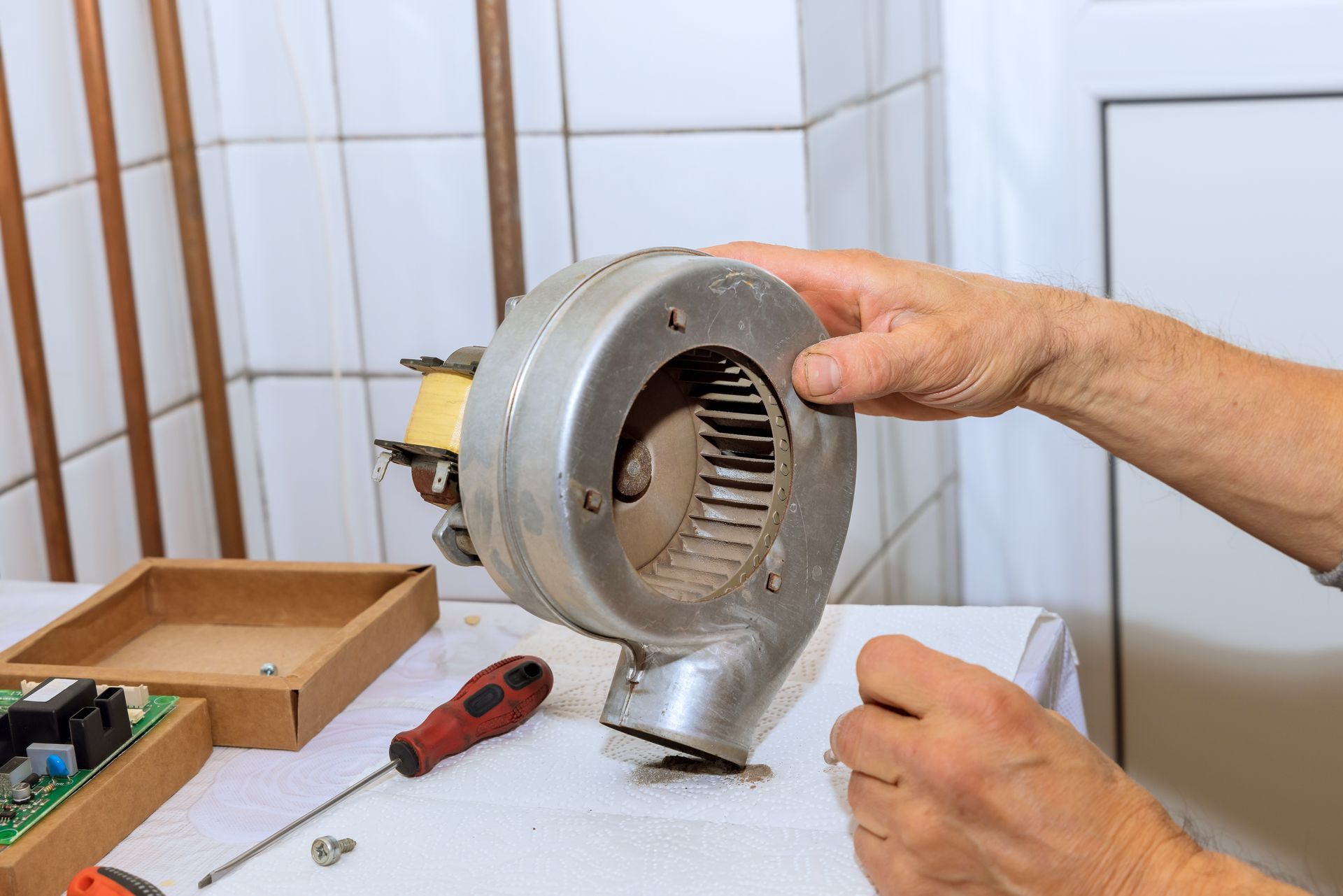 Person holding a blower fan during repair. A screwdriver, circuit board and other parts sit on a table.