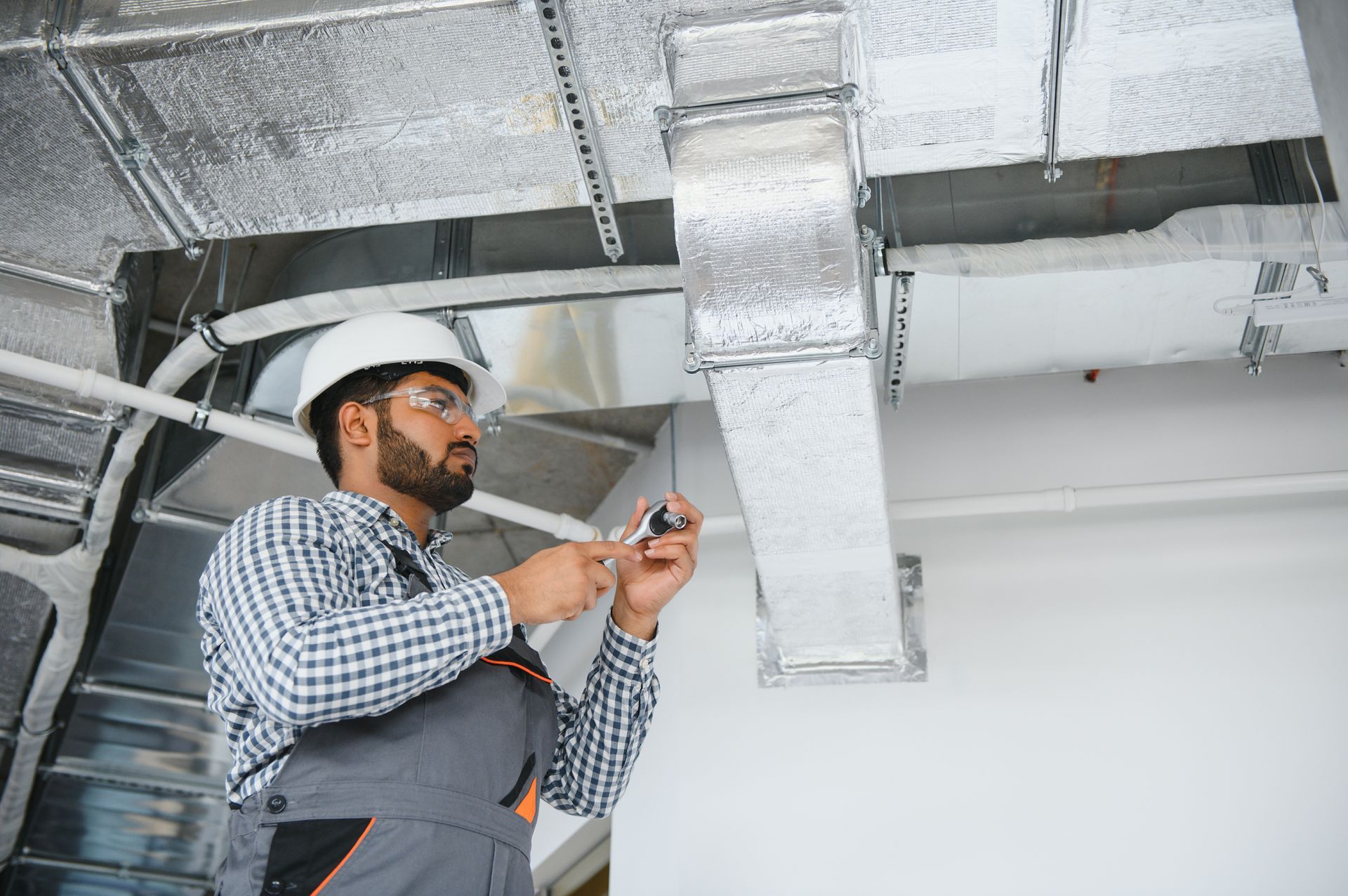 Construction worker inspecting ductwork, wearing hard hat, overalls, and safety glasses.