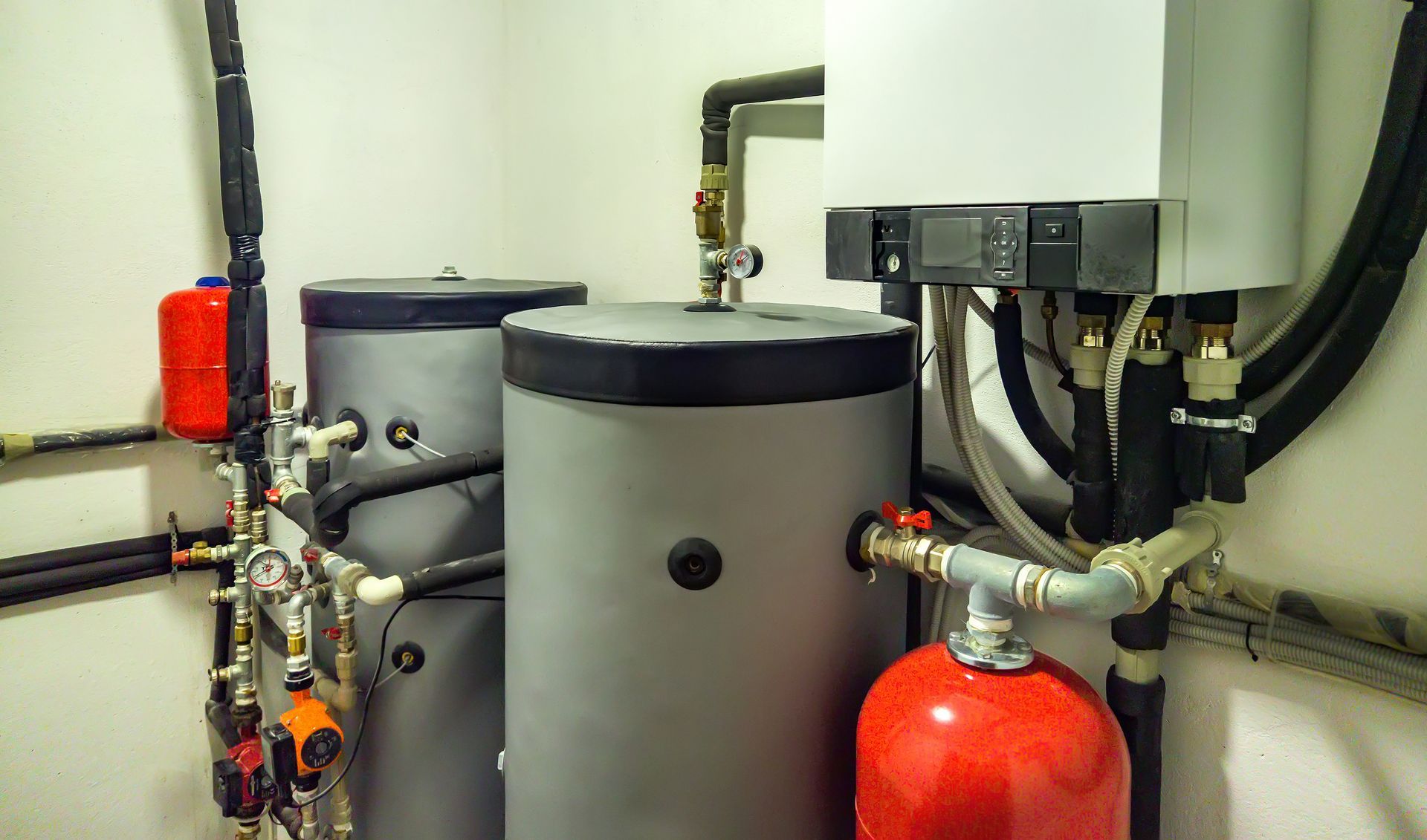 Water heater and related plumbing equipment in a white-walled room. Two gray tanks, red expansion tanks, and black pipes.