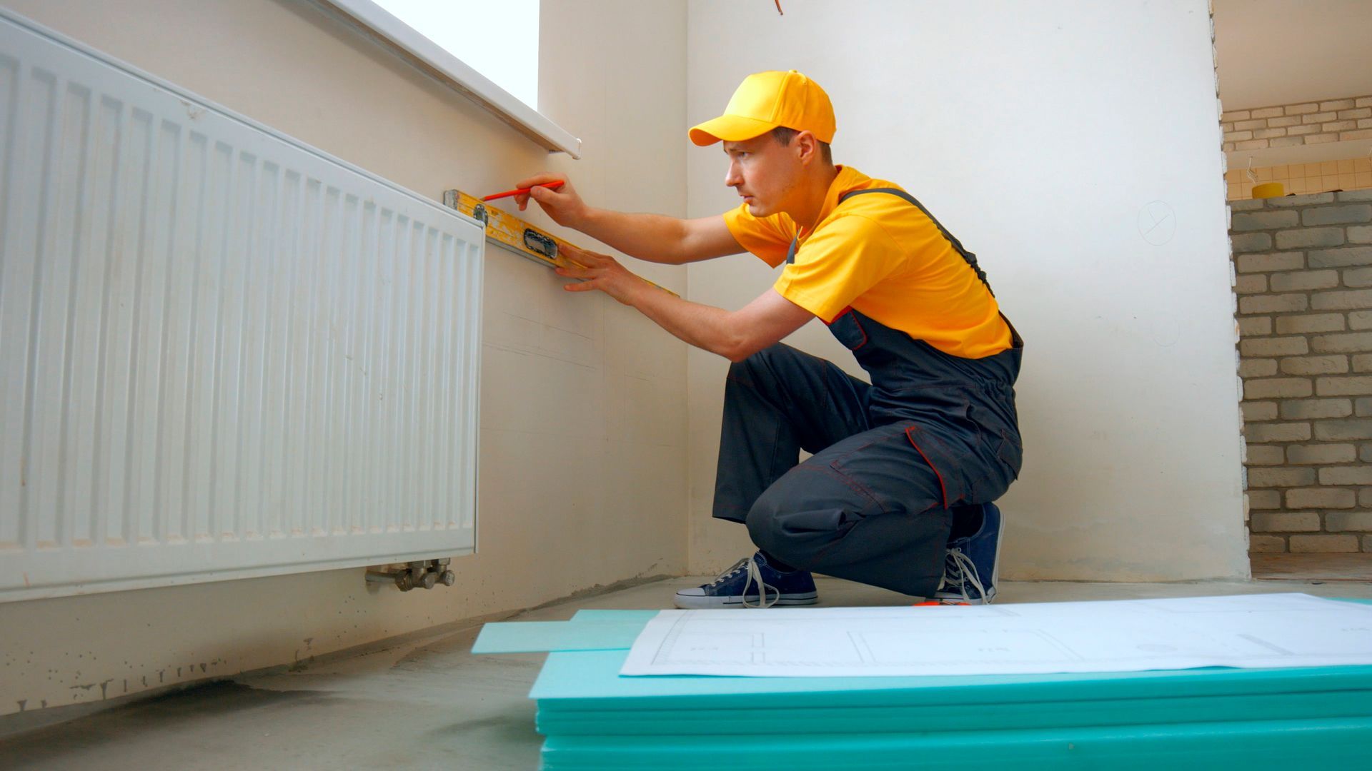 Man in yellow overalls and cap, kneeling, marking wall near a radiator.