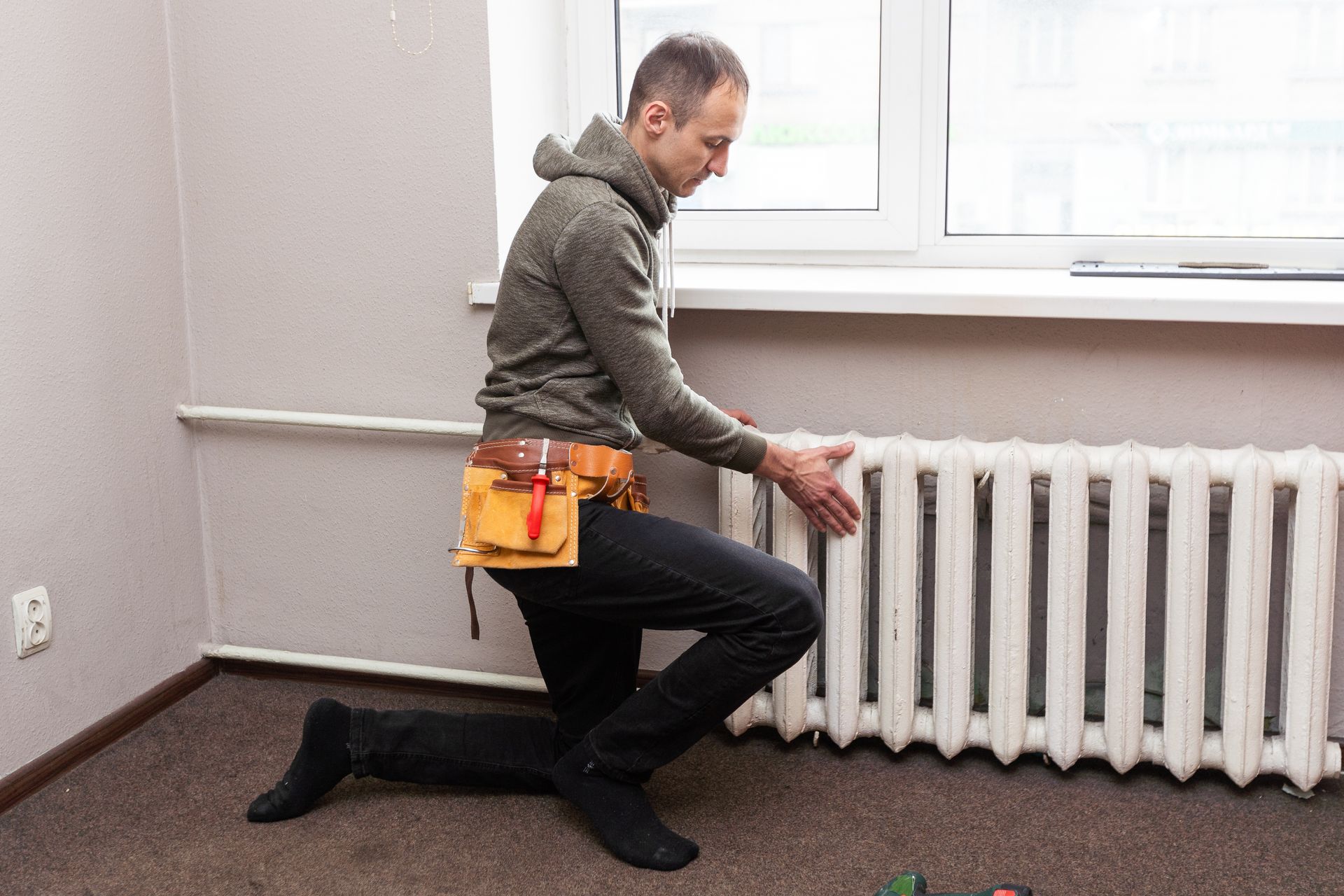 Man kneeling by a radiator, wearing a tool belt, inspecting it near a window.
