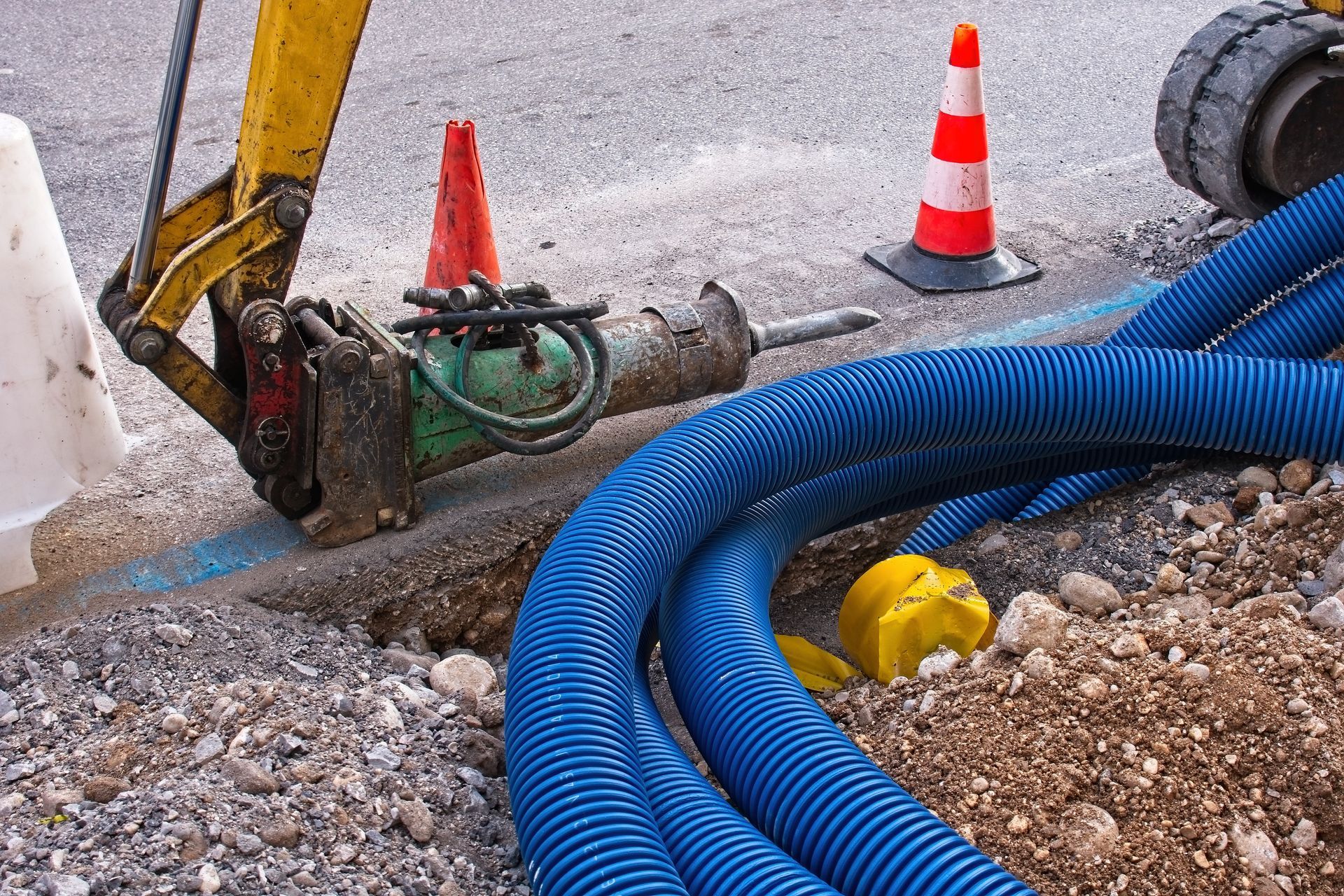 Construction site with blue corrugated pipes in a trench; a jackhammer and safety cones.