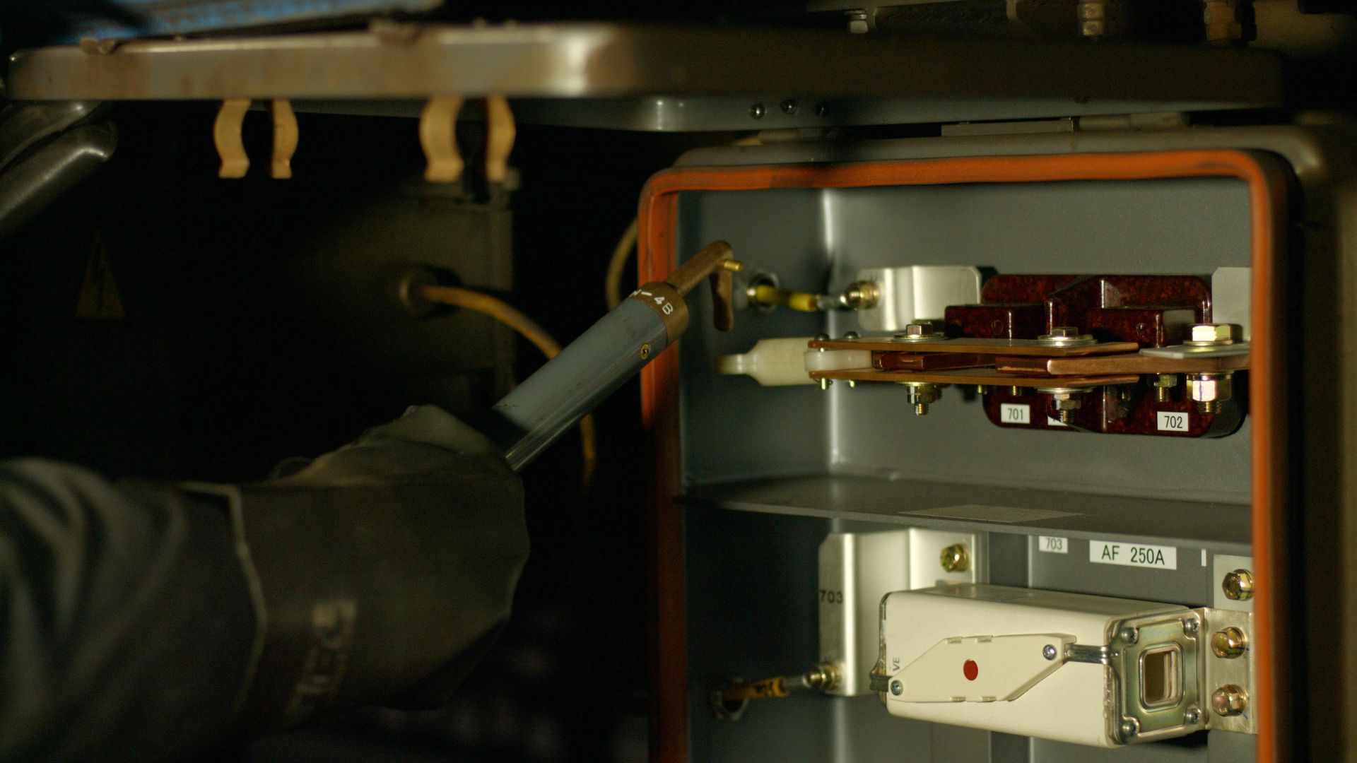 Gloved hands using a tool to work on electrical components inside a metal box, likely for repair or maintenance.