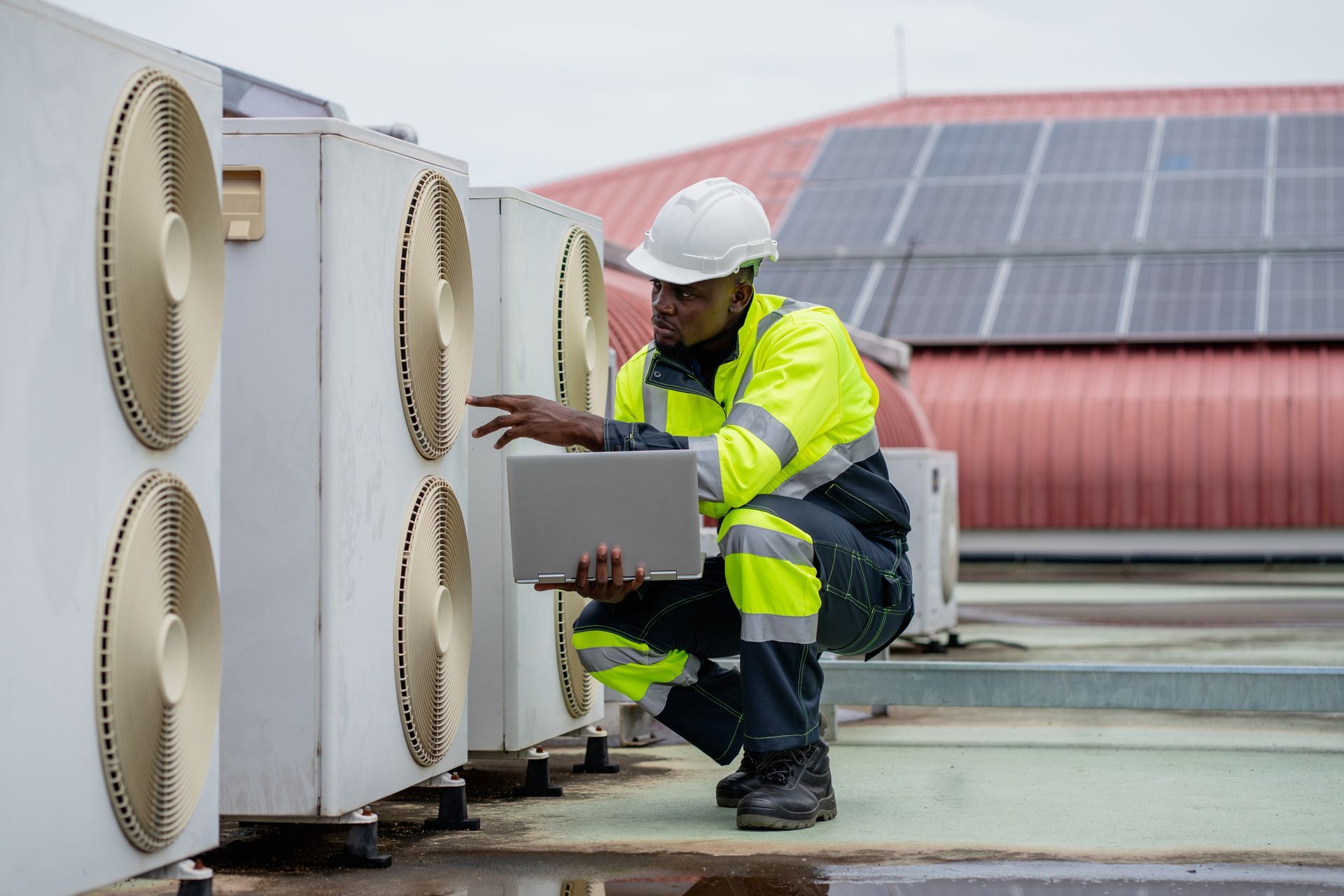 A Black HVAC technician in safety gear, inspecting rooftop cooling units with a laptop.