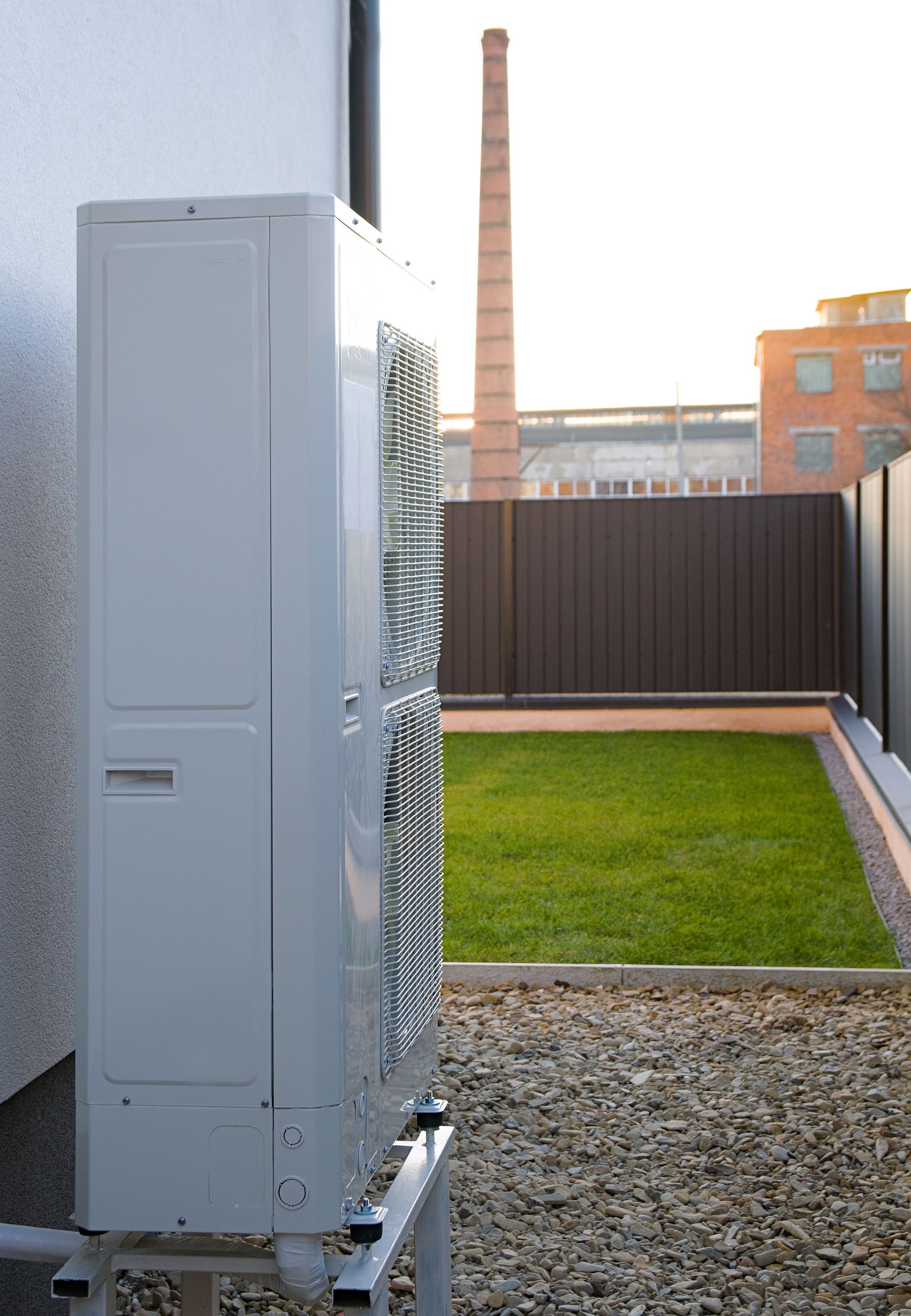 Air conditioner unit on a building's side, with a grassy yard and a factory chimney in the background.