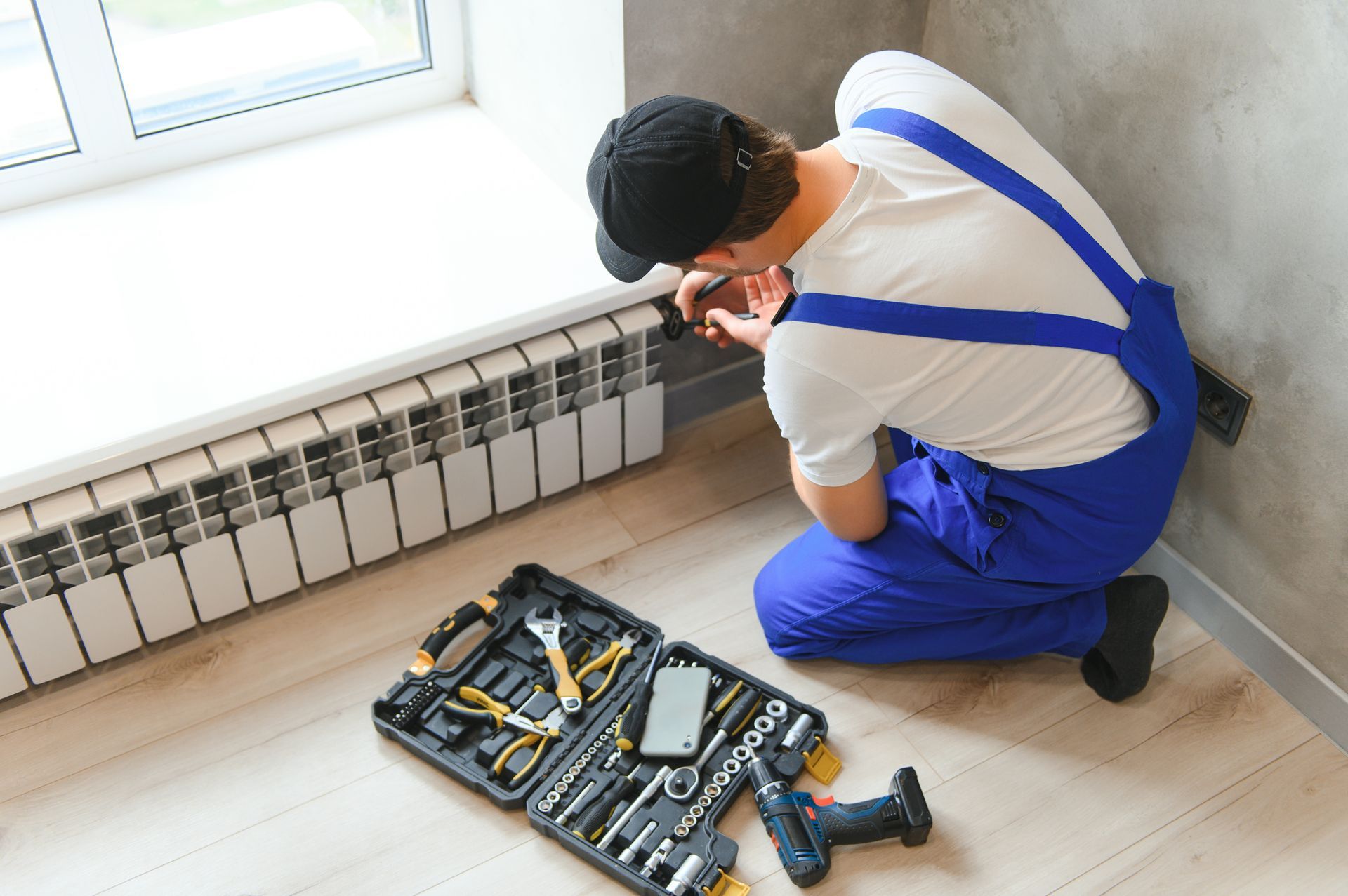 Man in blue overalls installing a radiator, tools nearby in a case, by a window.