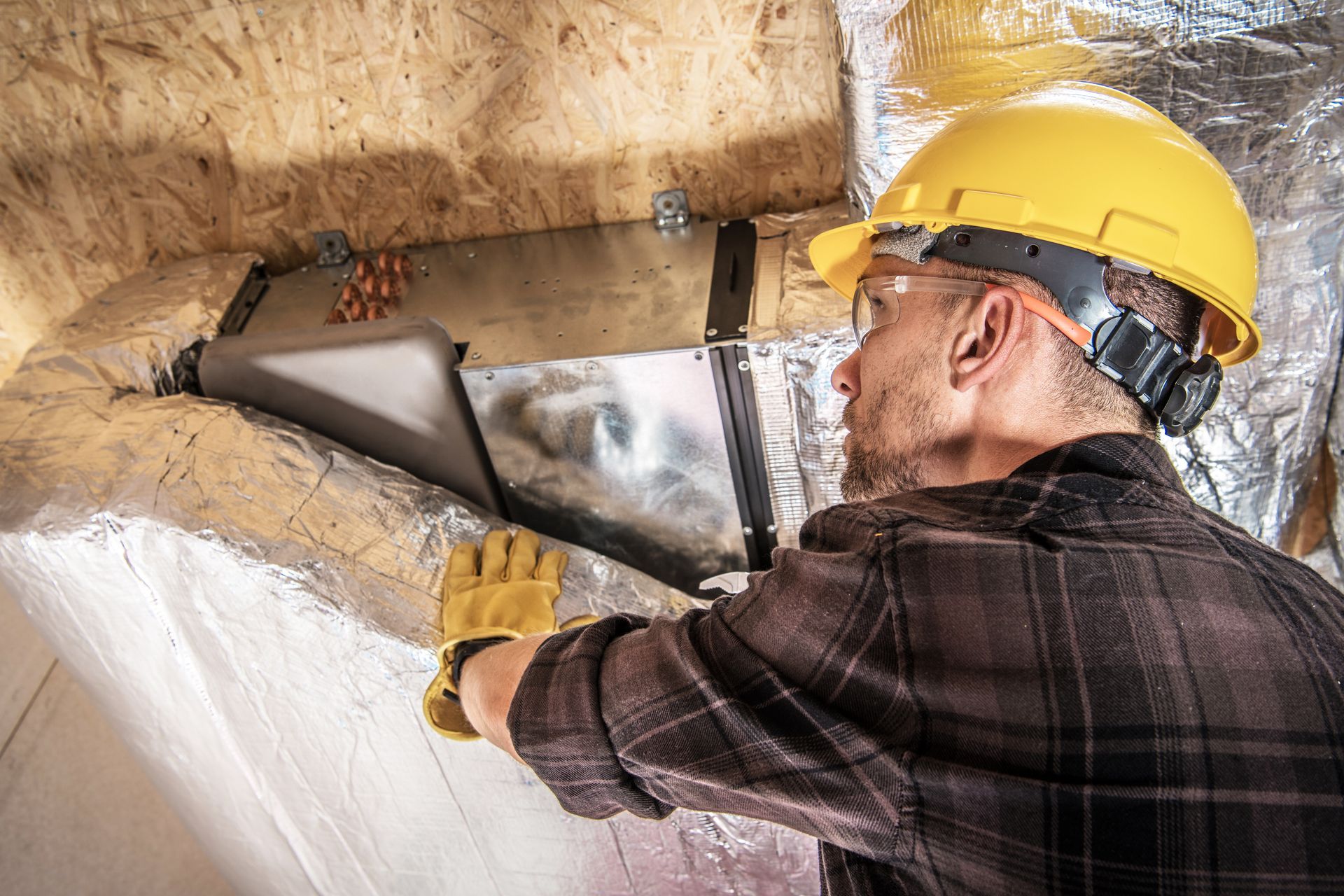 A worker in a hard hat and gloves installs HVAC ductwork in an attic.