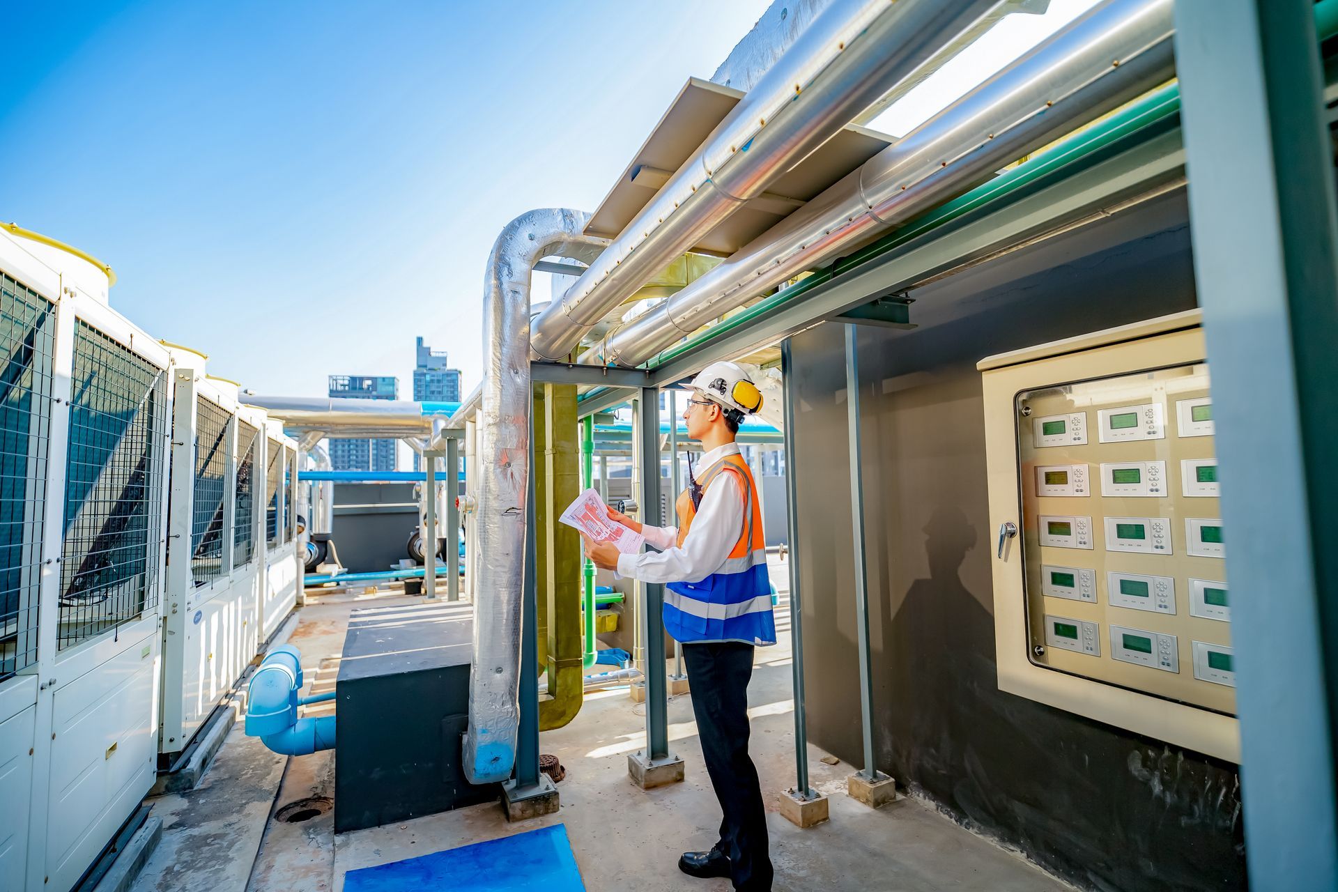 Engineer in hard hat and vest examines pipes on a building rooftop, blue sky background.