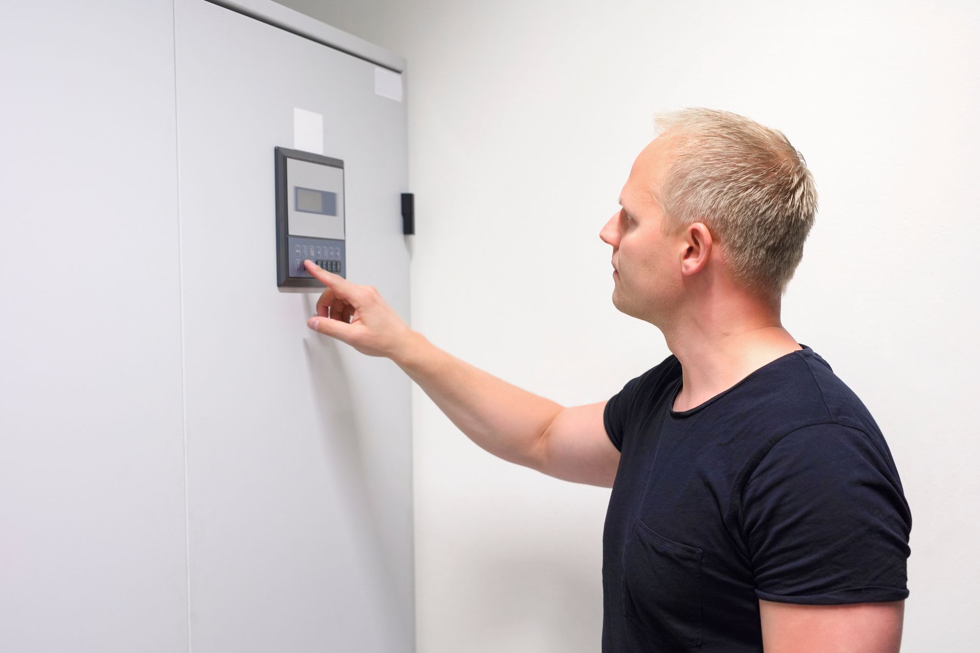 Man with fair skin presses a button on a gray control panel mounted on a white wall.