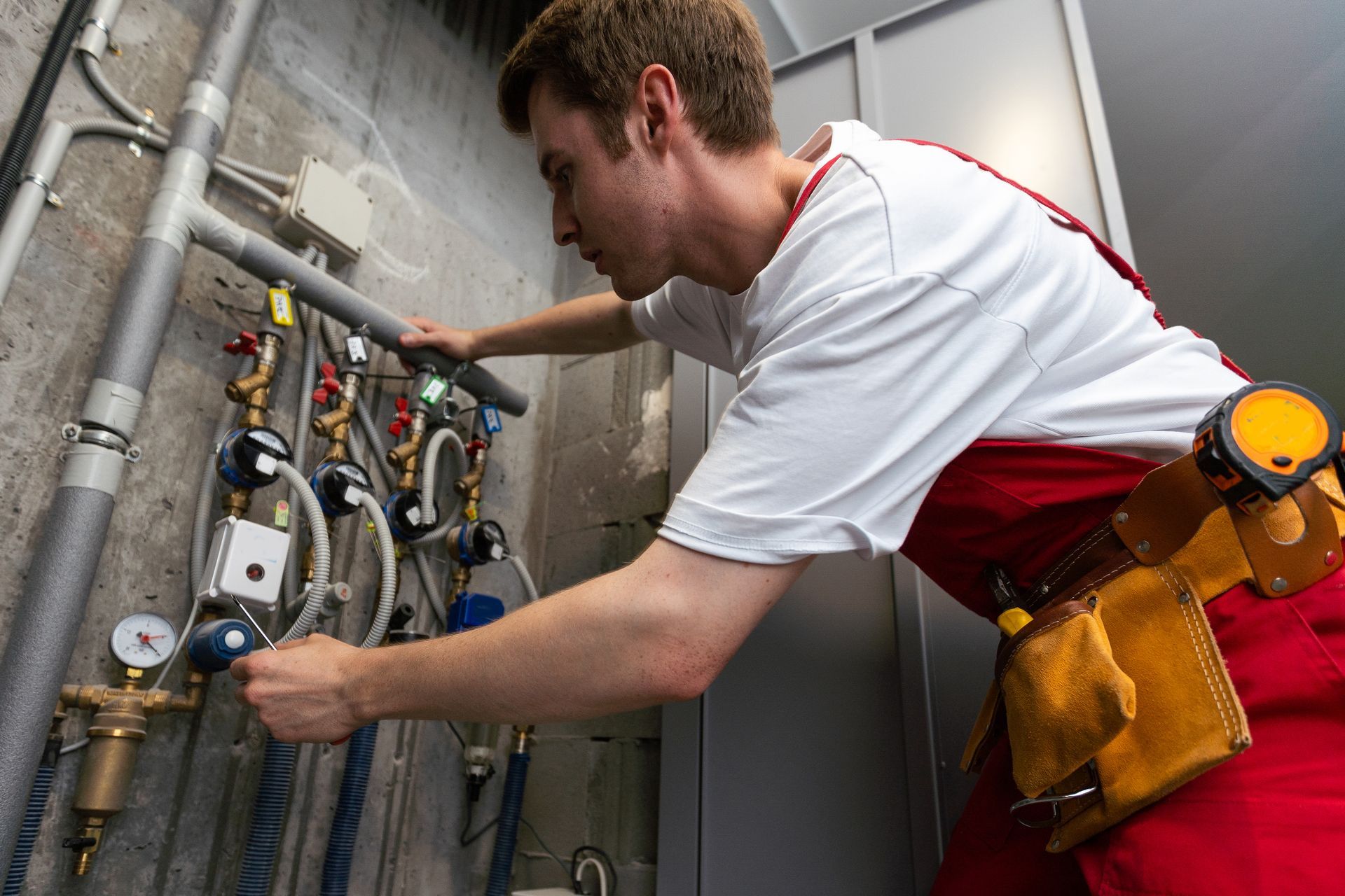A plumber in red overalls, works on pipes and gauges in an industrial setting.
