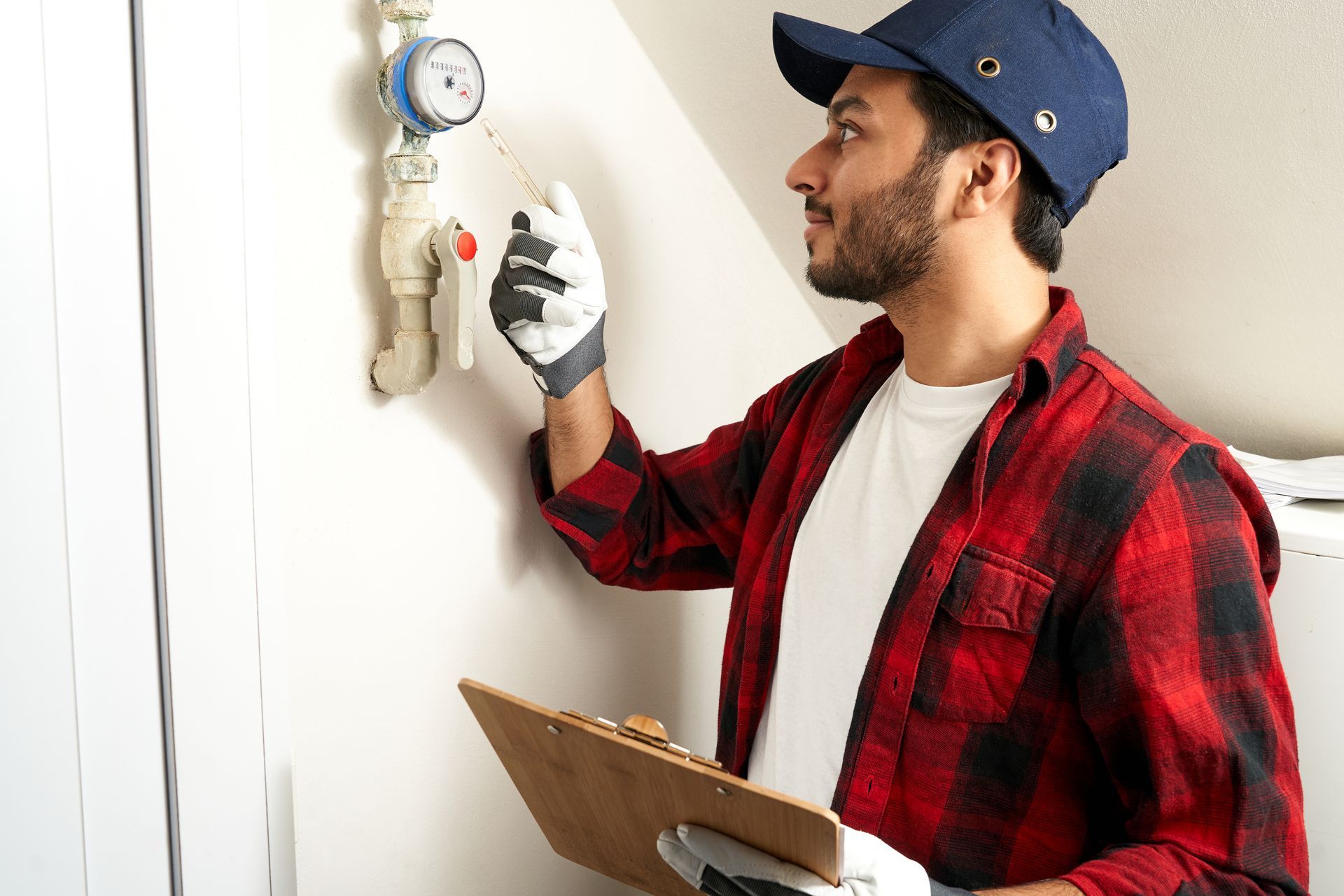 Plumber in red plaid shirt and cap inspects a pipe and water meter in a room, holding a clipboard.