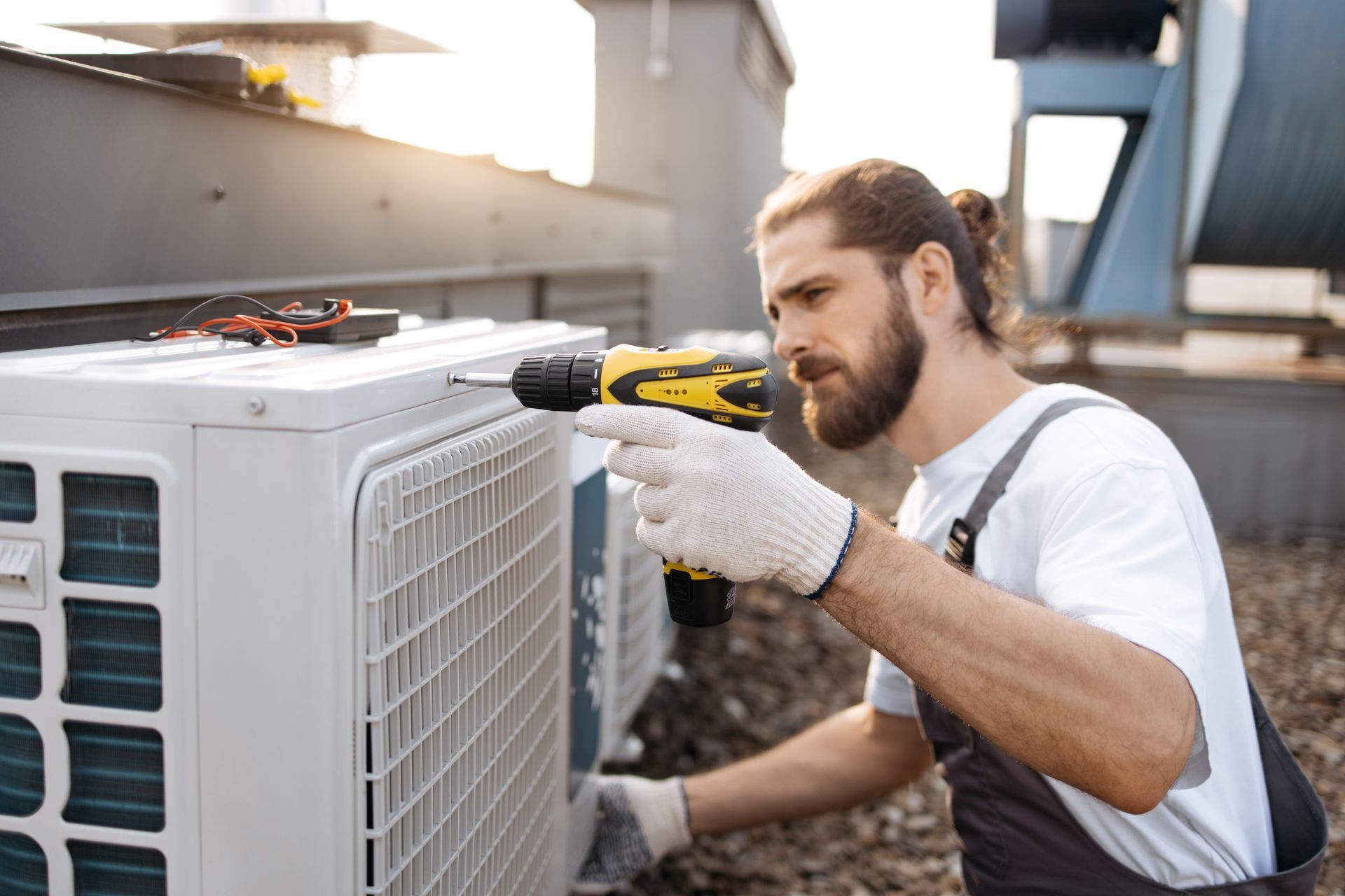 HVAC technician in overalls, fixing an AC unit on a rooftop, using a drill.