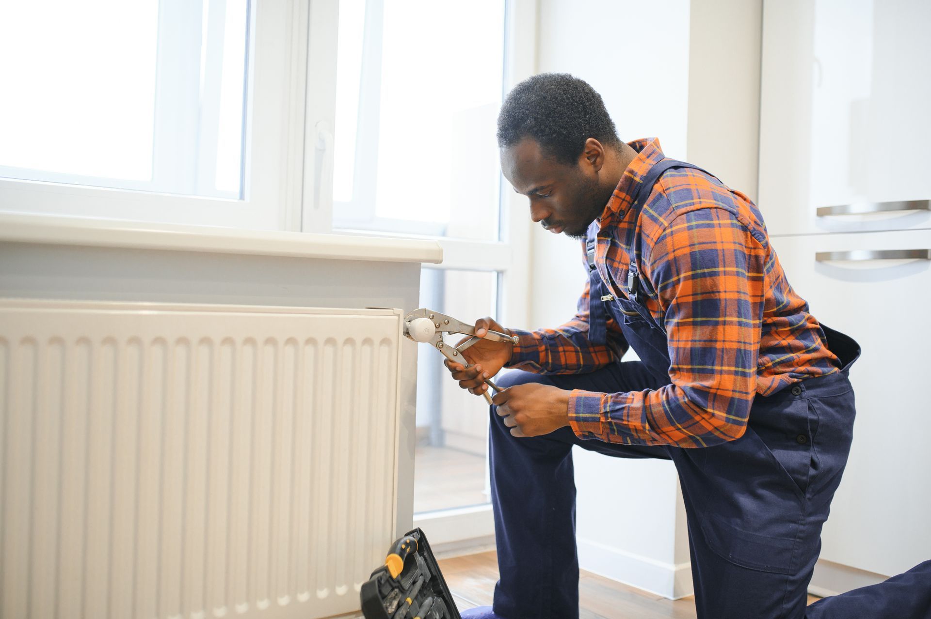 A Black man in overalls kneels, using pliers on a white radiator next to a window.