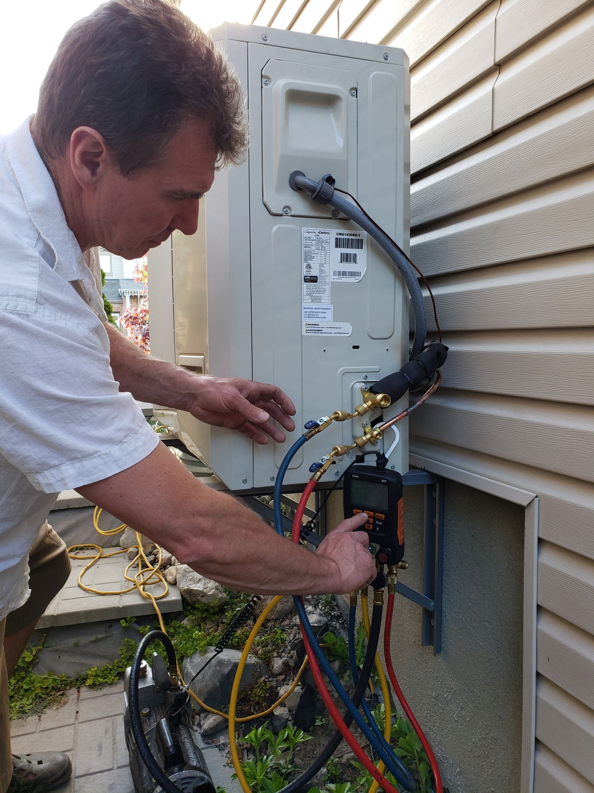 Man servicing outdoor air conditioner, using gauges. Against a wall.