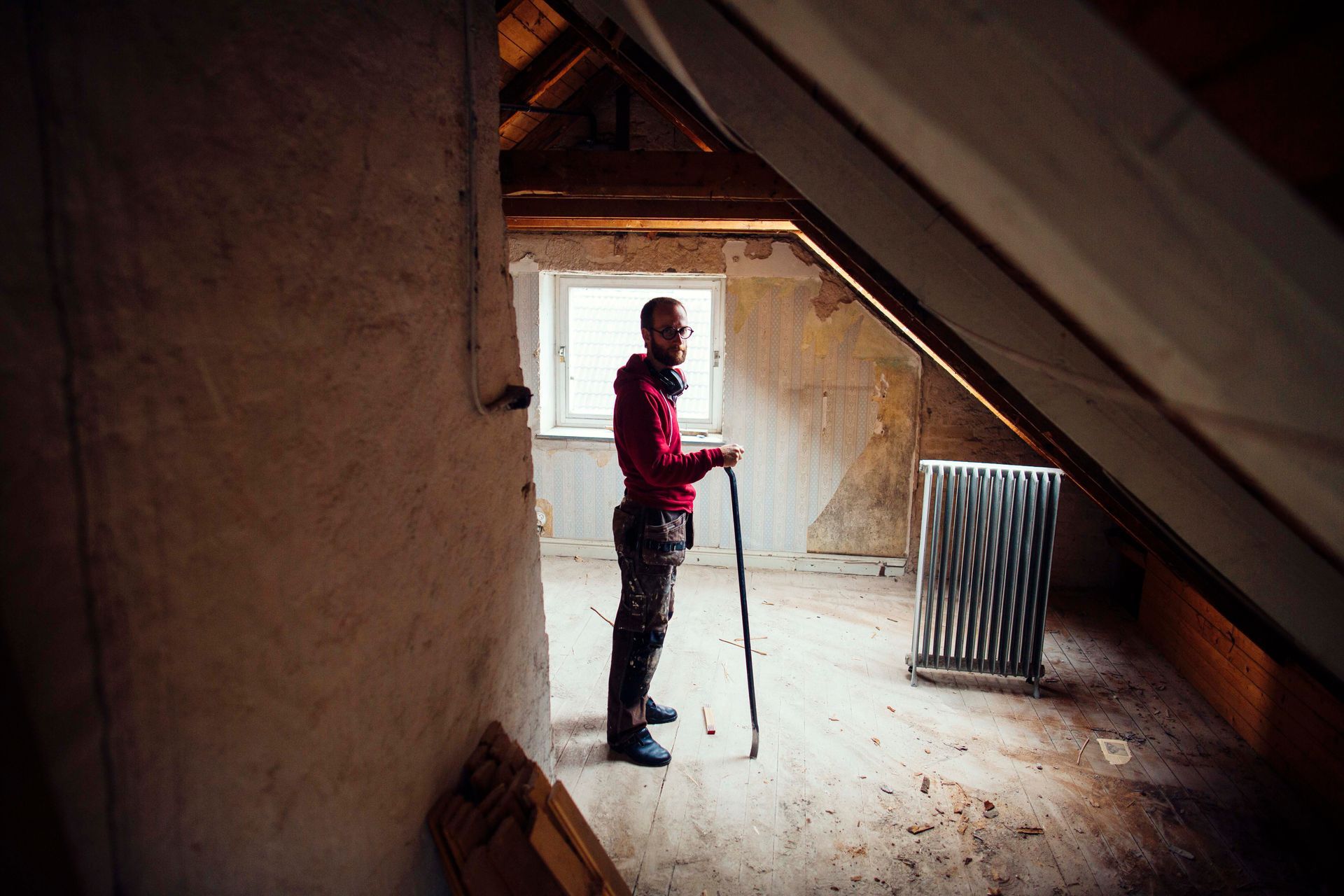 Man in red shirt and patterned pants stands in an attic, holding a cane.
