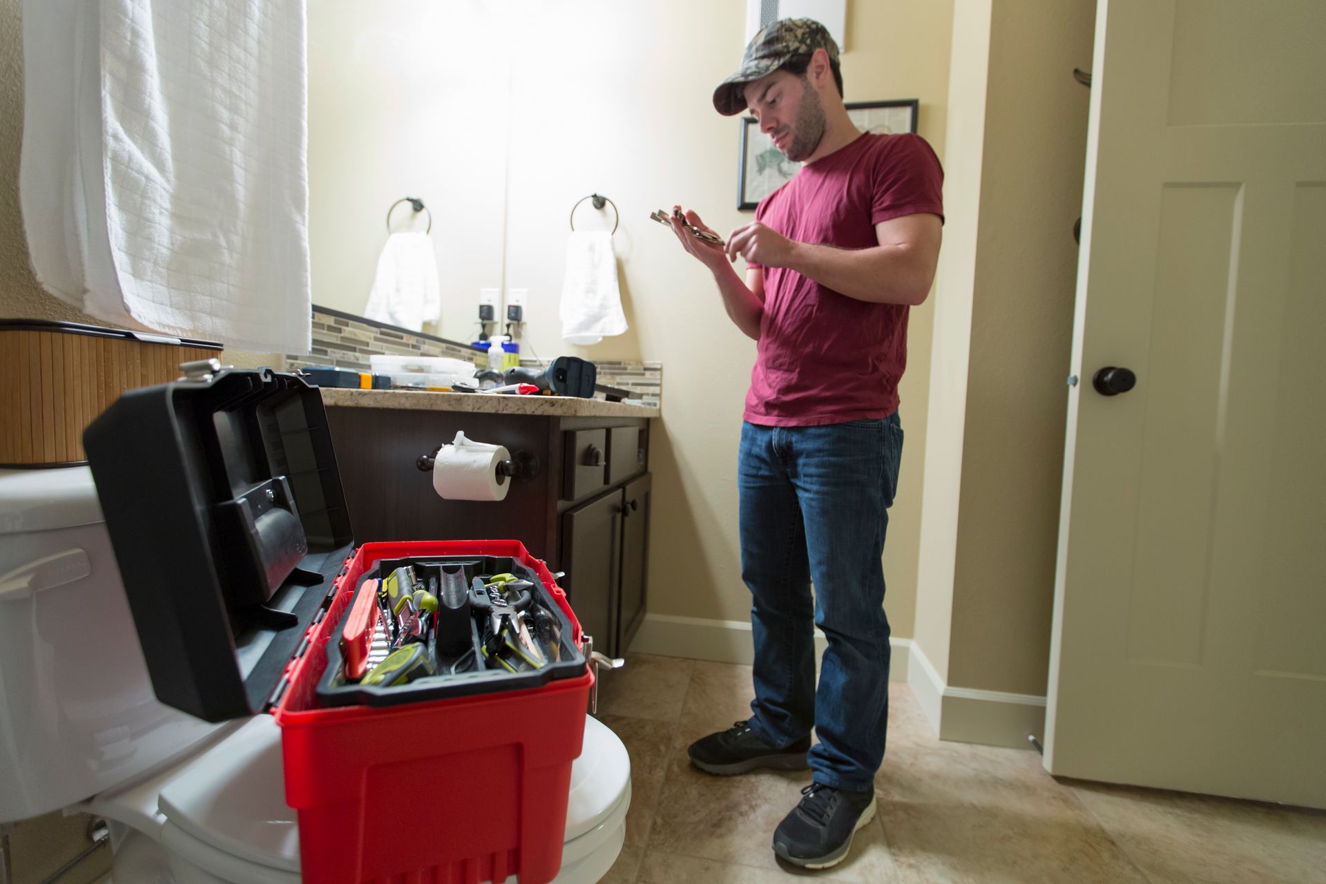 Man in bathroom, examining tools from an open red toolbox on a toilet, next to a sink and mirror.