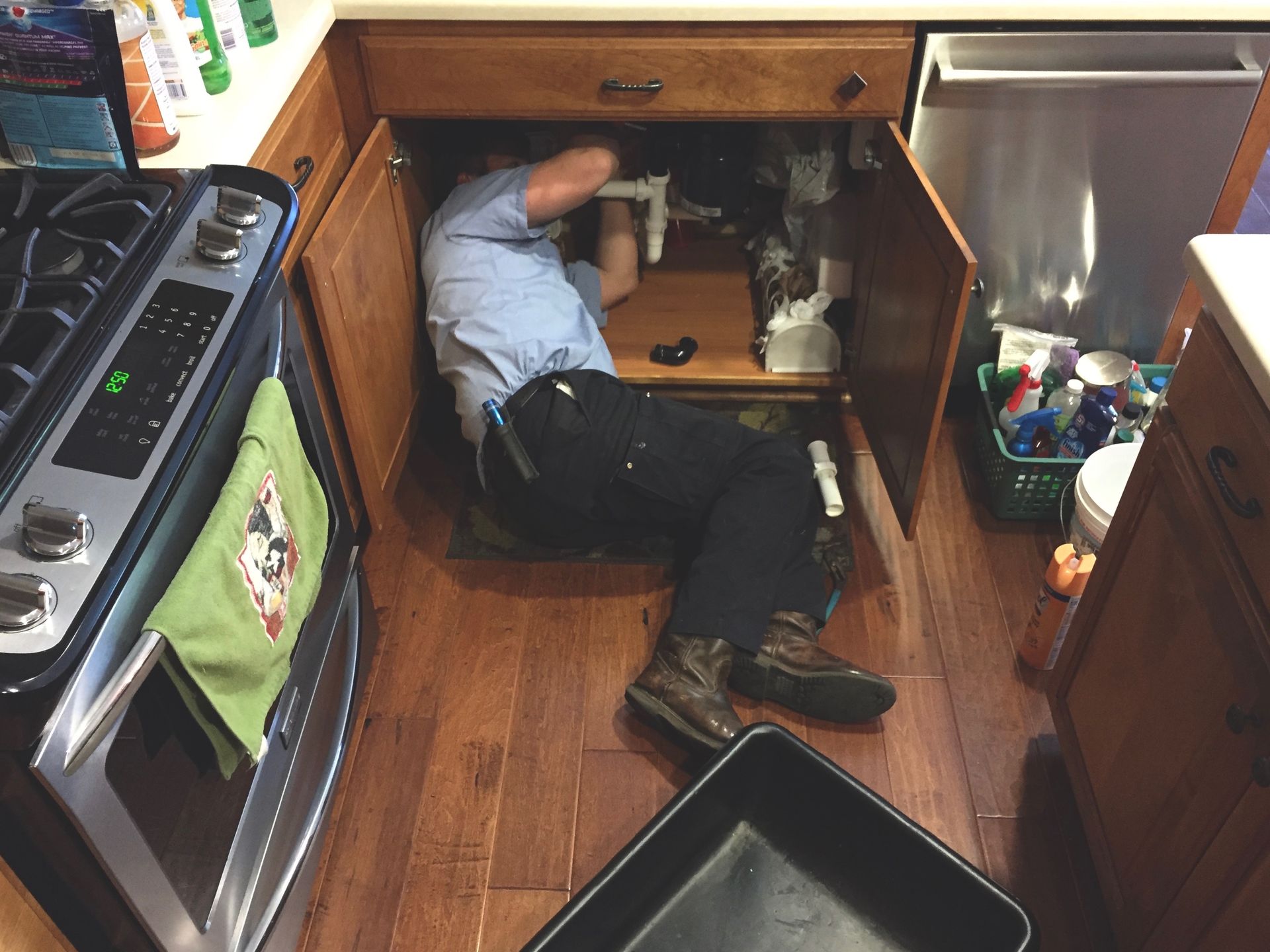A plumber works under a kitchen sink with tools. He's wearing boots and dark pants.