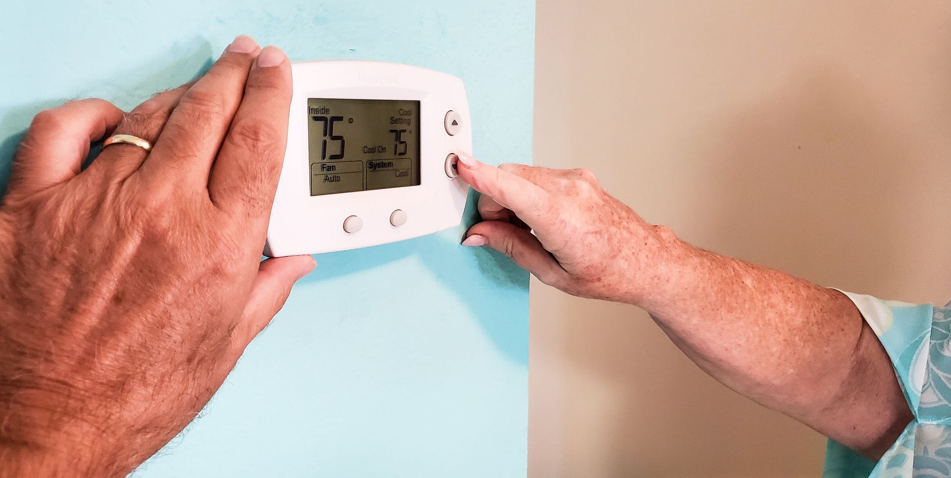 Two people adjusting a thermostat on a light blue wall. One hand holds the thermostat while the other presses a button.