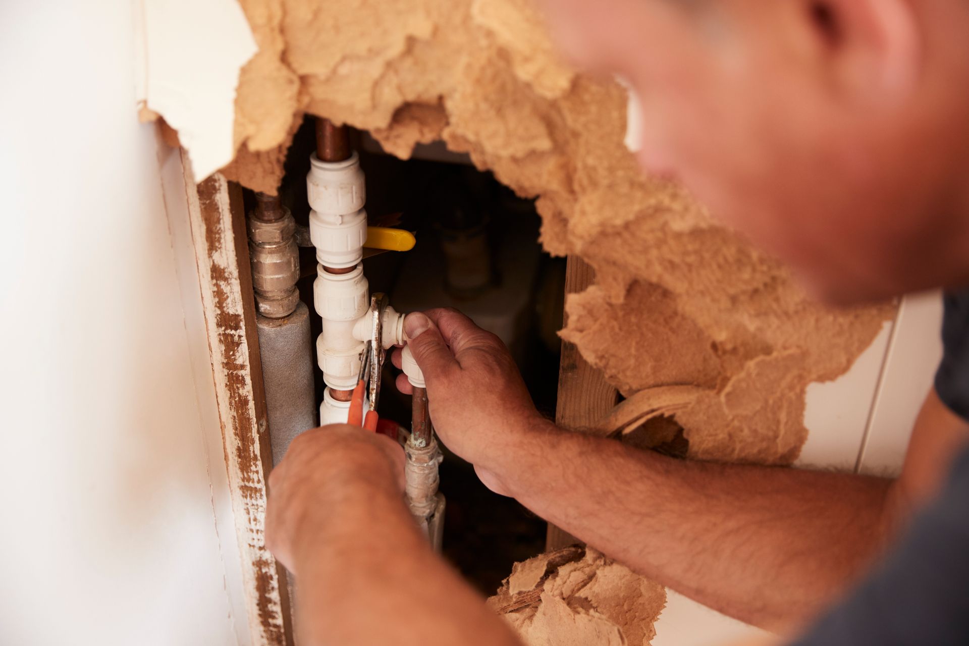 Plumber repairs pipes inside a wall, using pliers. Brown insulation is exposed around a hole.