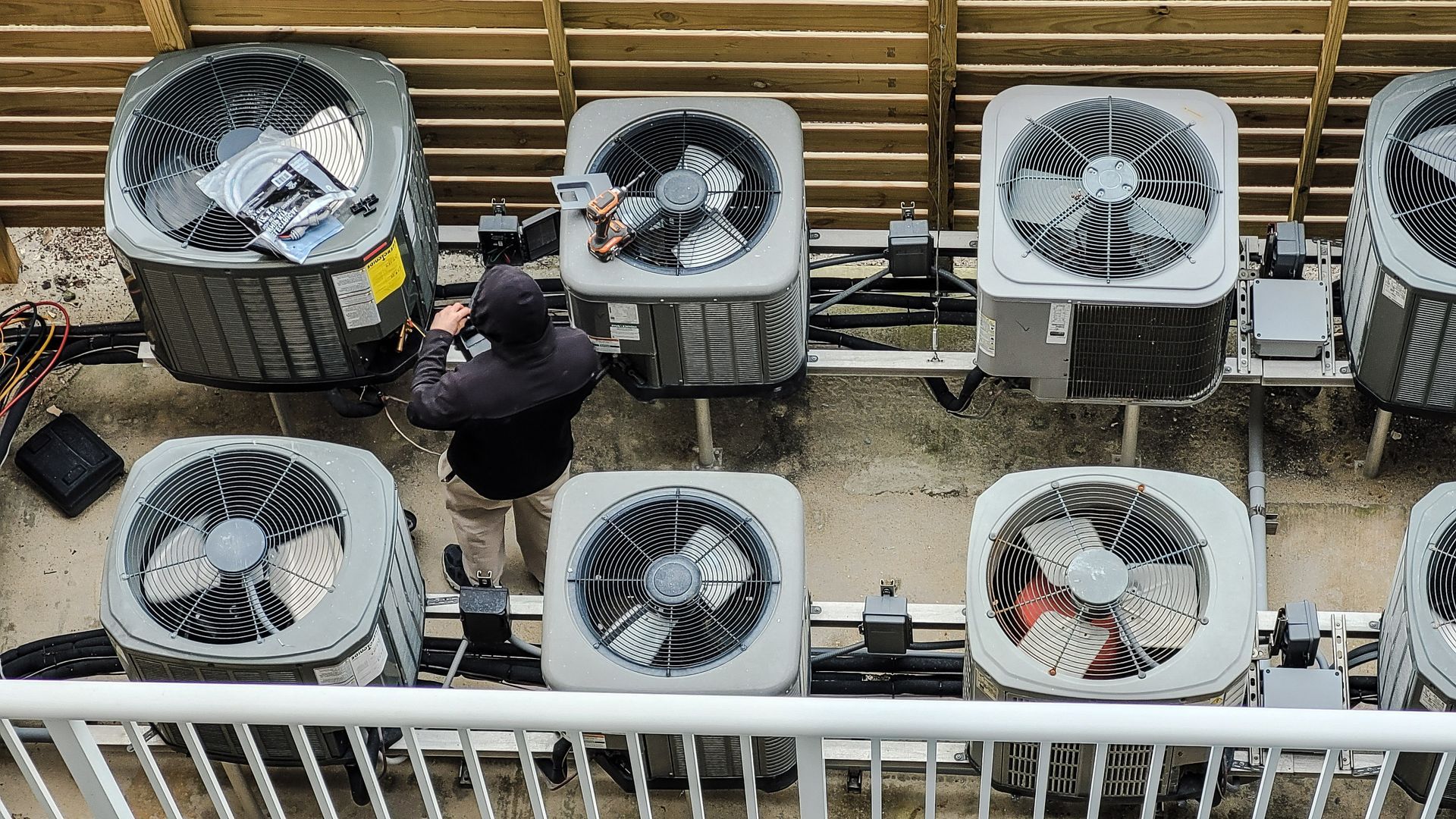 A person repairs an air conditioning unit among several other units on a rooftop.