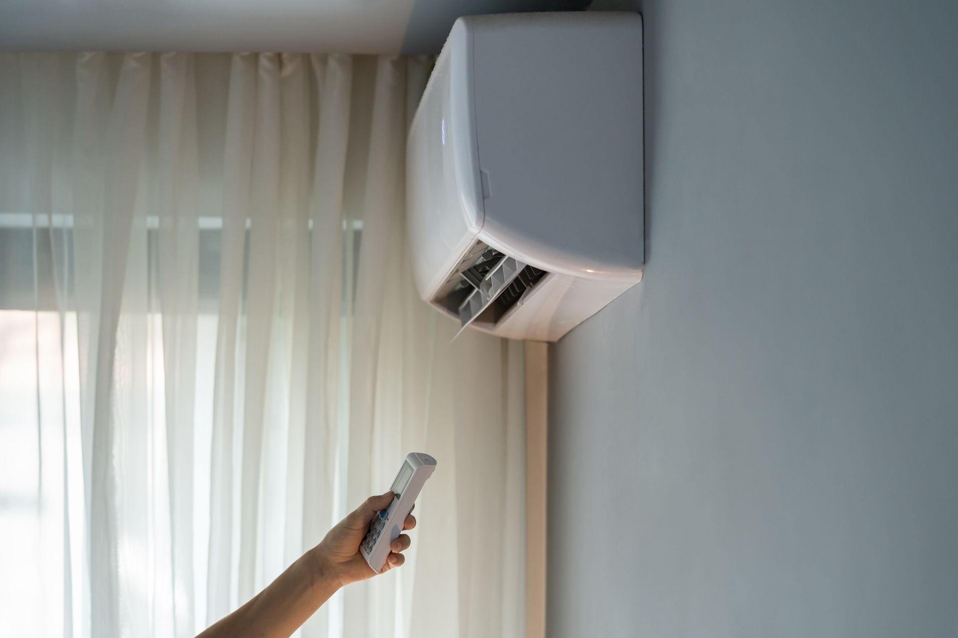 Hand holding a remote, pointing at a wall-mounted air conditioner near a sheer curtain.