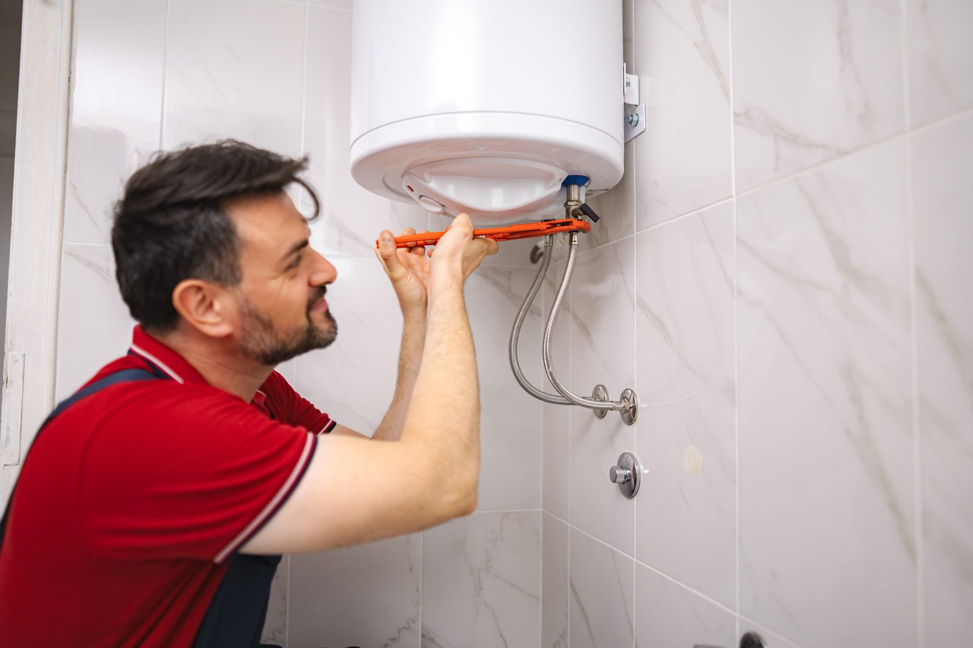 A plumber in a red shirt is fixing a water heater with a wrench in a bathroom.