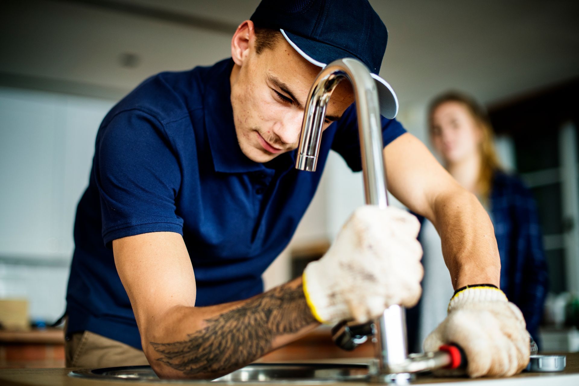 Plumber in blue shirt and cap, fixing kitchen faucet with a wrench, woman in background.