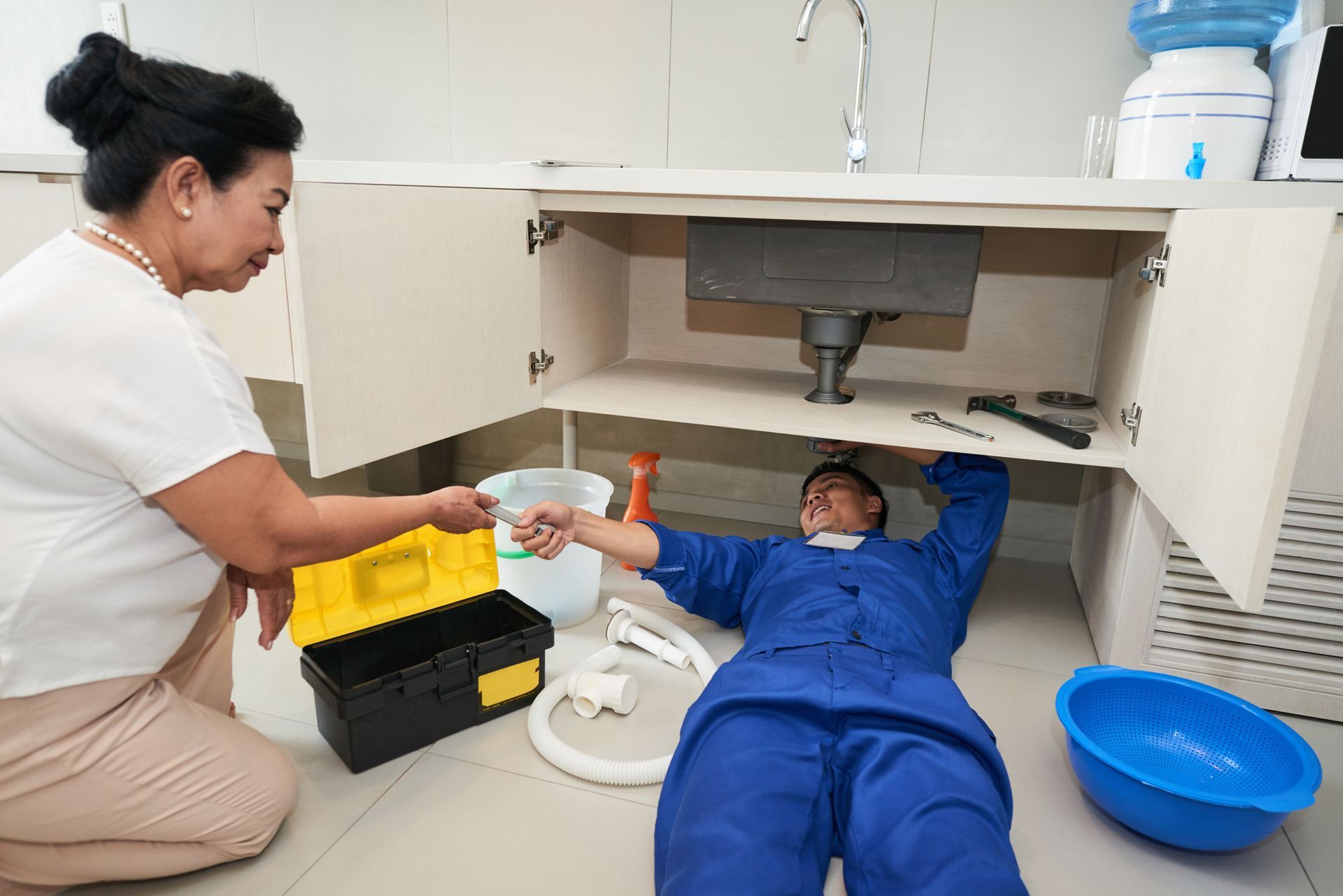 Woman helping a plumber working under a kitchen sink; white cabinets, tools visible.