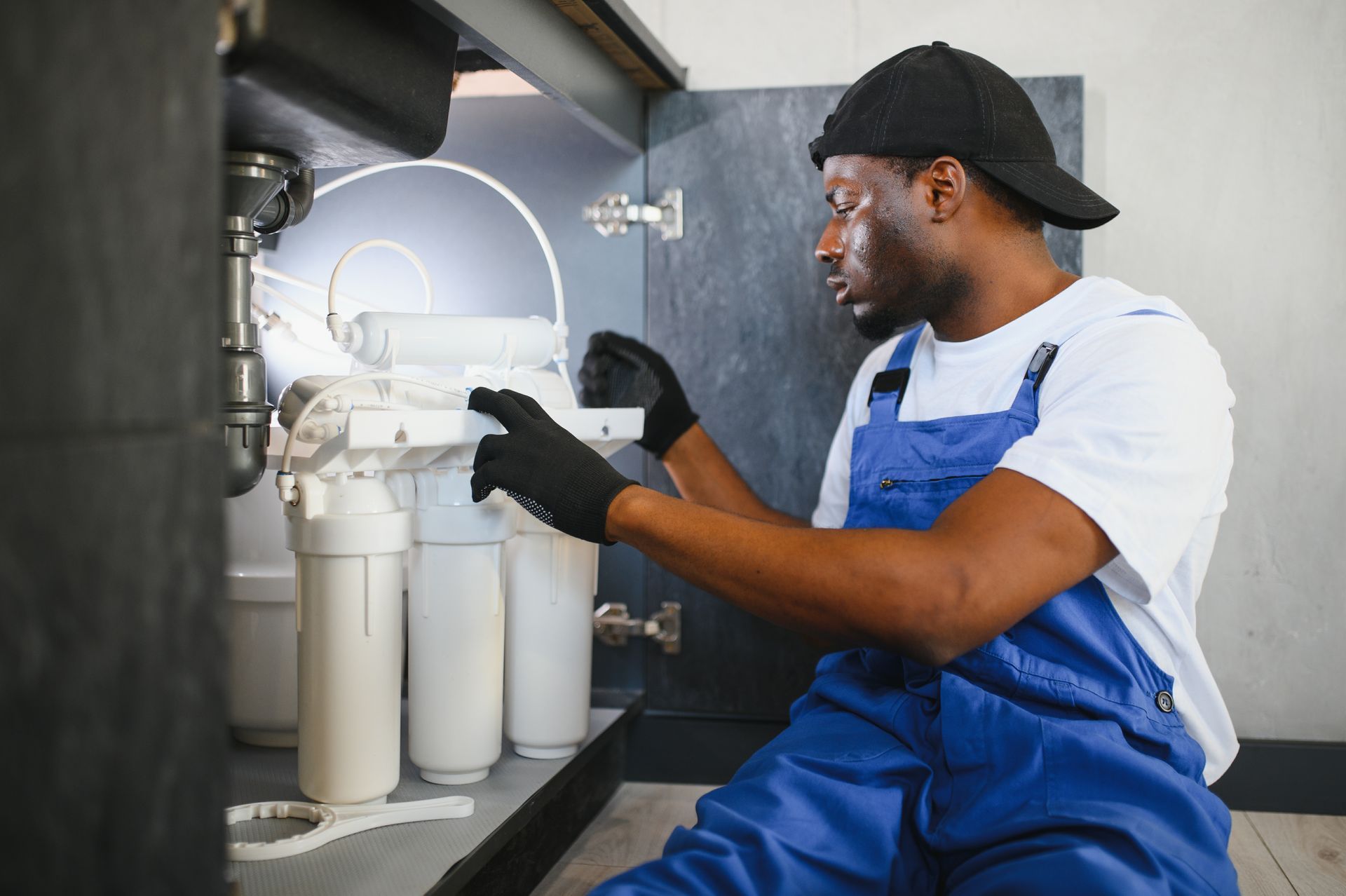 A Black plumber in blue overalls installs a water filter under a sink.