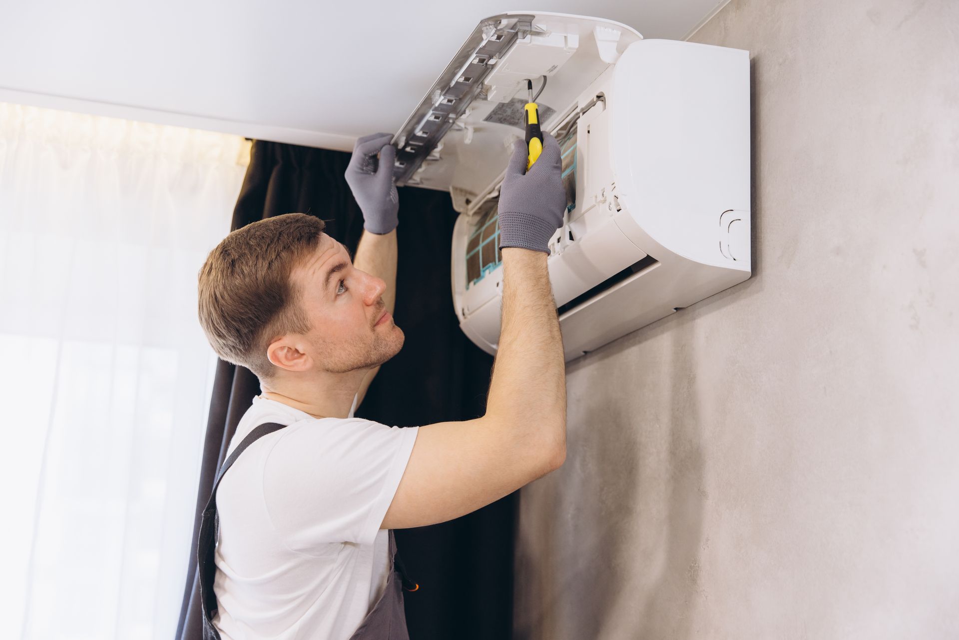 Man in gray gloves repairs wall-mounted AC unit with a screwdriver.