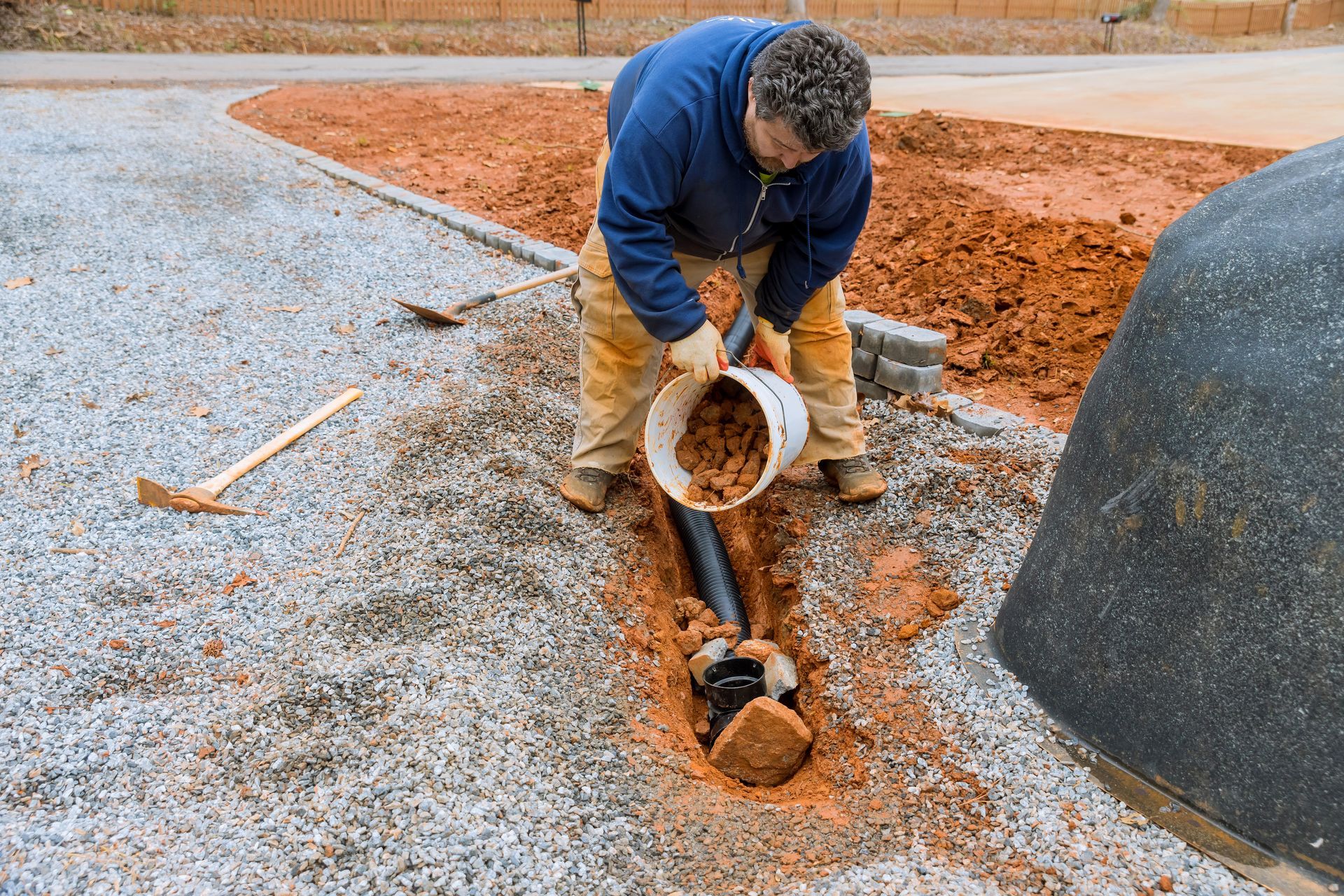 Man in blue shirt pours red soil into a trench containing a black pipe, gravel path and red clay background.