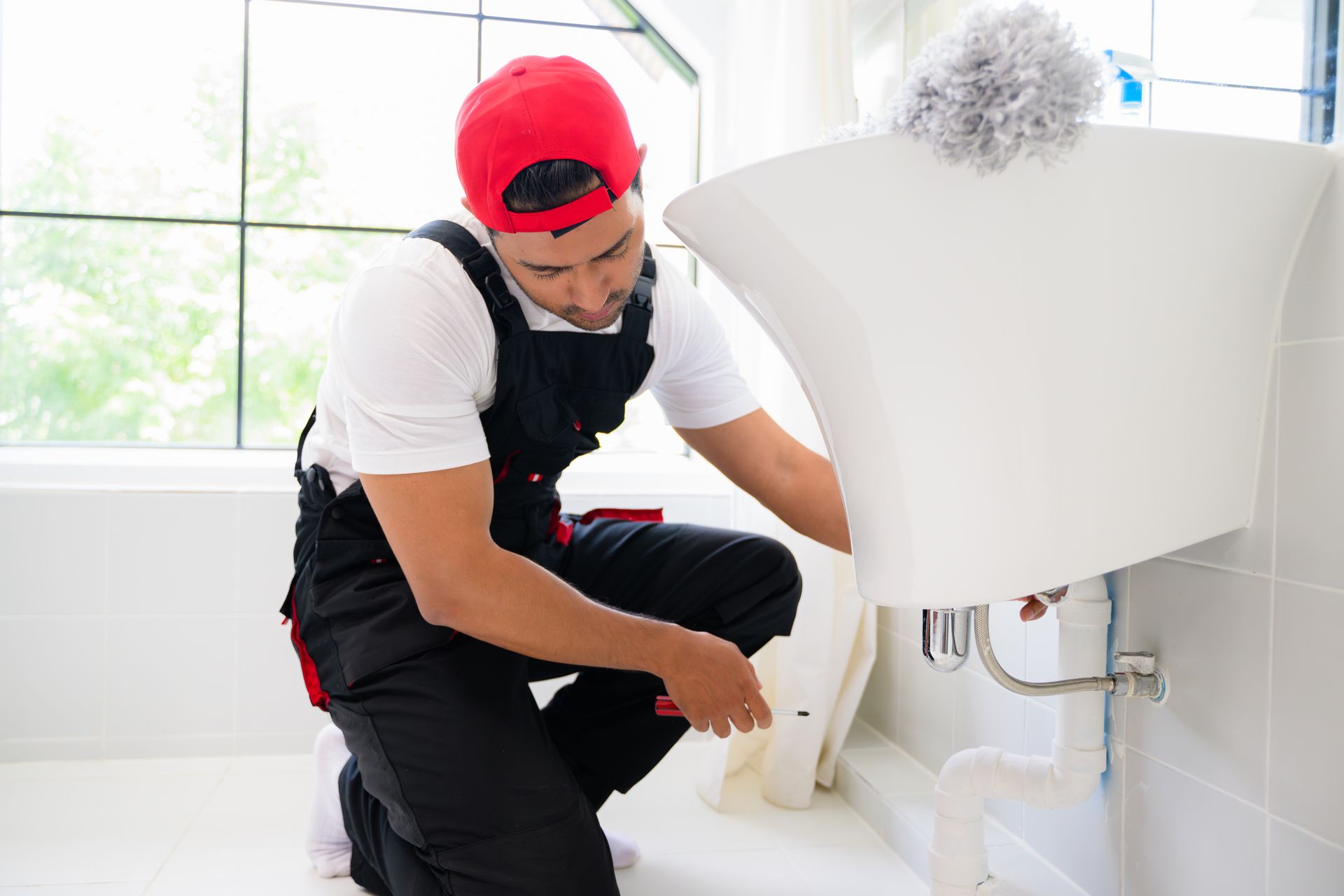 Plumber in red cap and overalls kneels by a white sink, repairing plumbing in a bright bathroom.