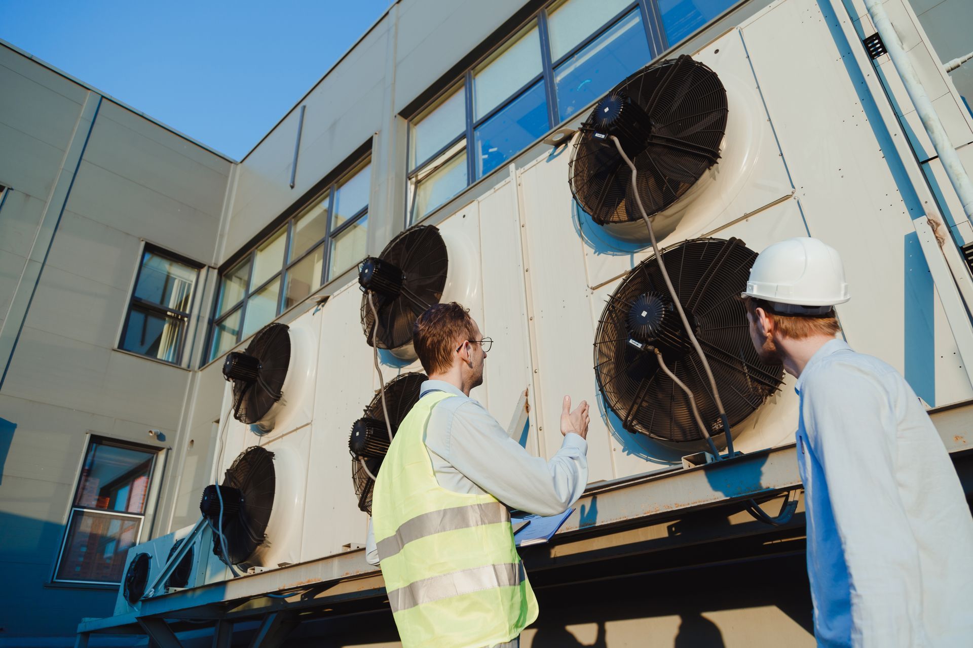 Two men in safety gear examine HVAC units outside a building.
