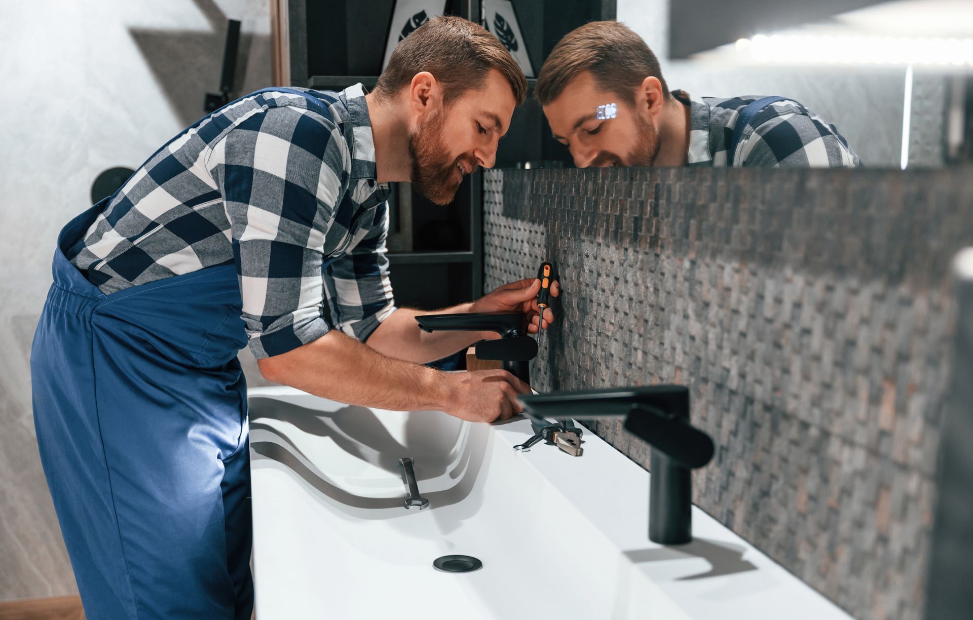 A plumber installing a black faucet in a modern bathroom, reflected in the mirror.