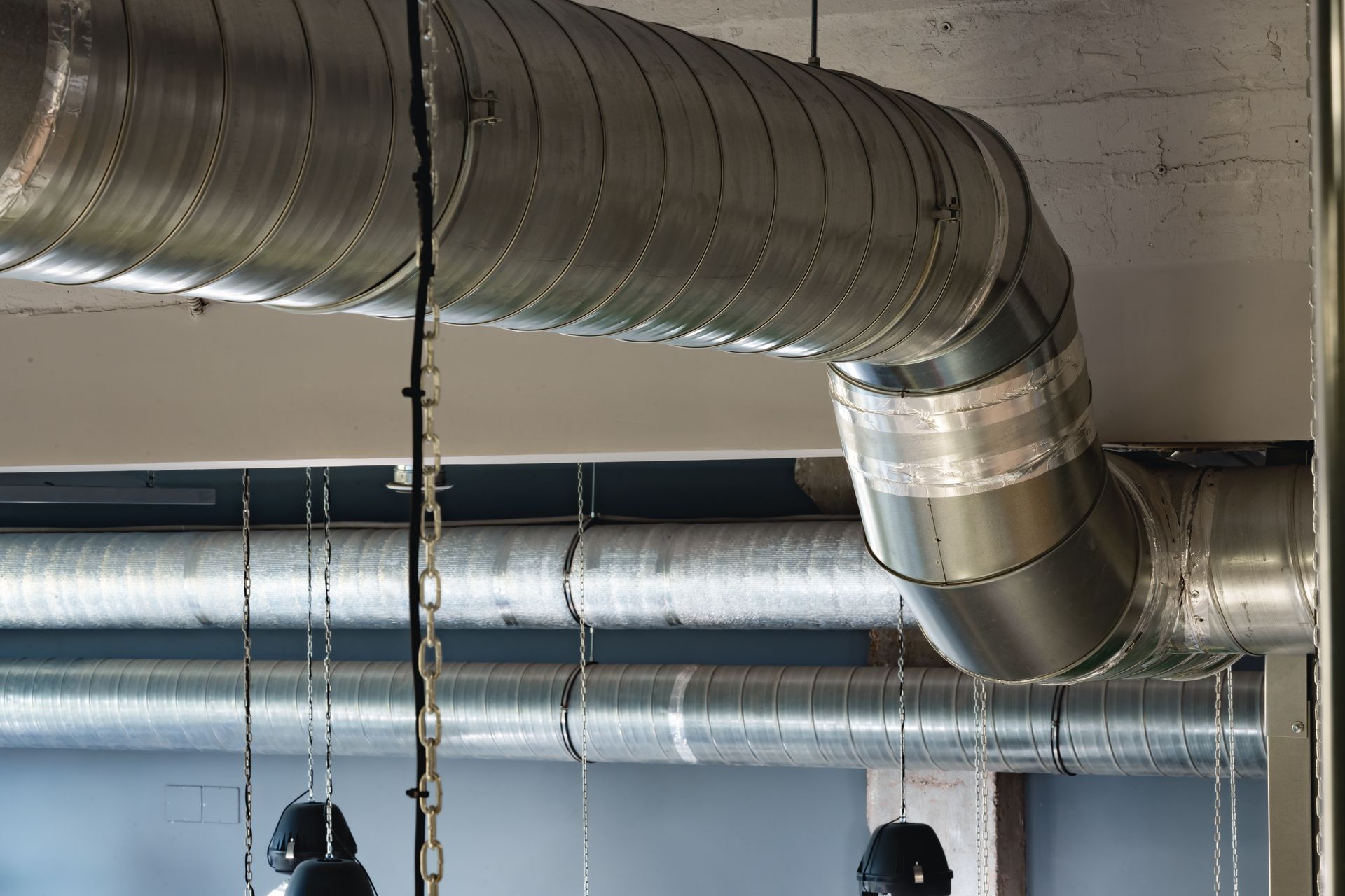 Metal ductwork, curved, with visible seams, in an industrial setting with hanging chains and fixtures.
