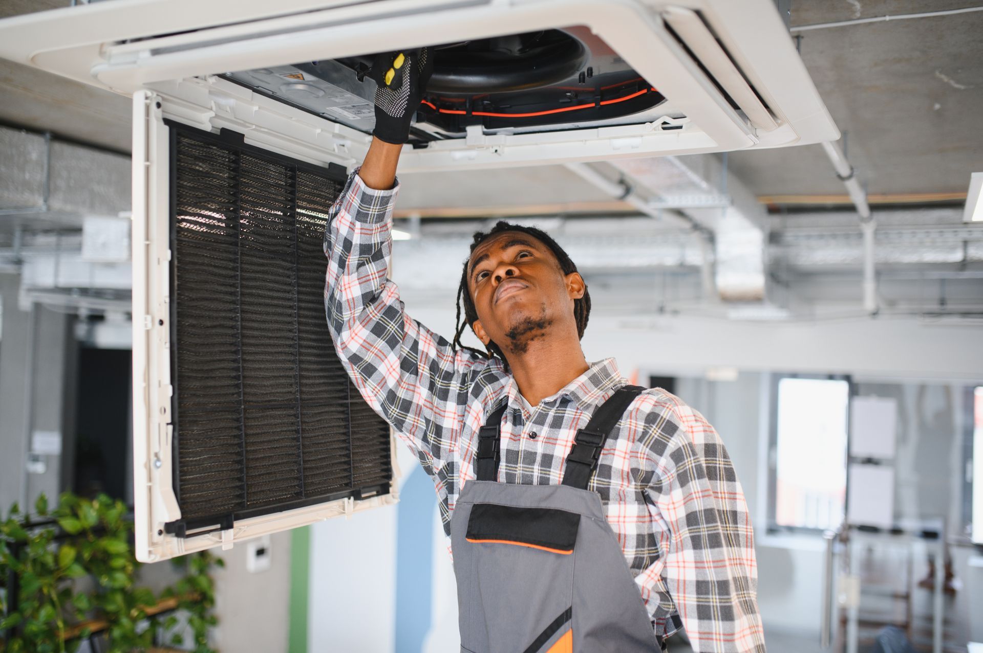 Black HVAC technician repairs ceiling air conditioner. He wears overalls, a plaid shirt, and gloves in a modern office.