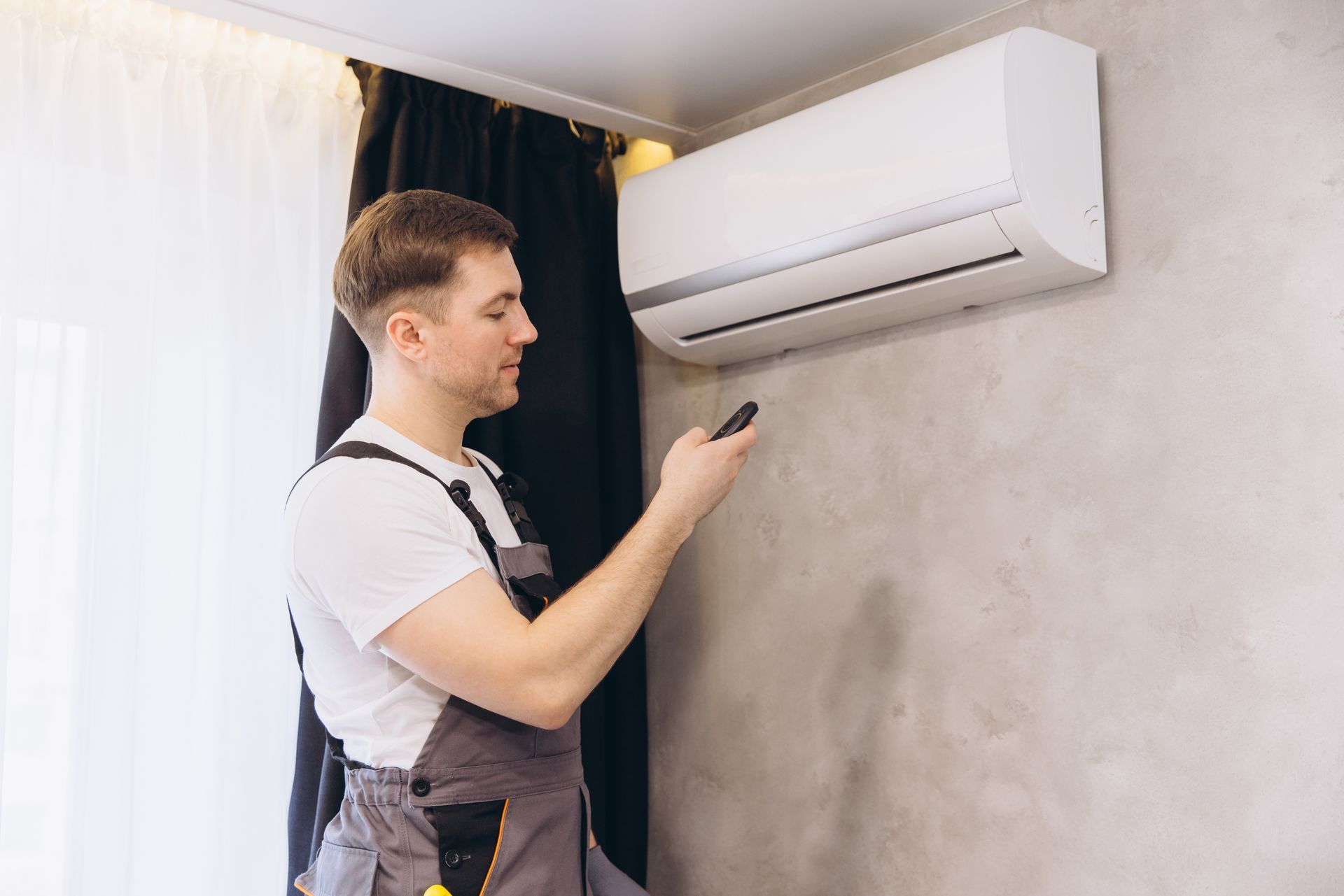 Man in work clothes using a remote to adjust an air conditioner mounted on a wall.
