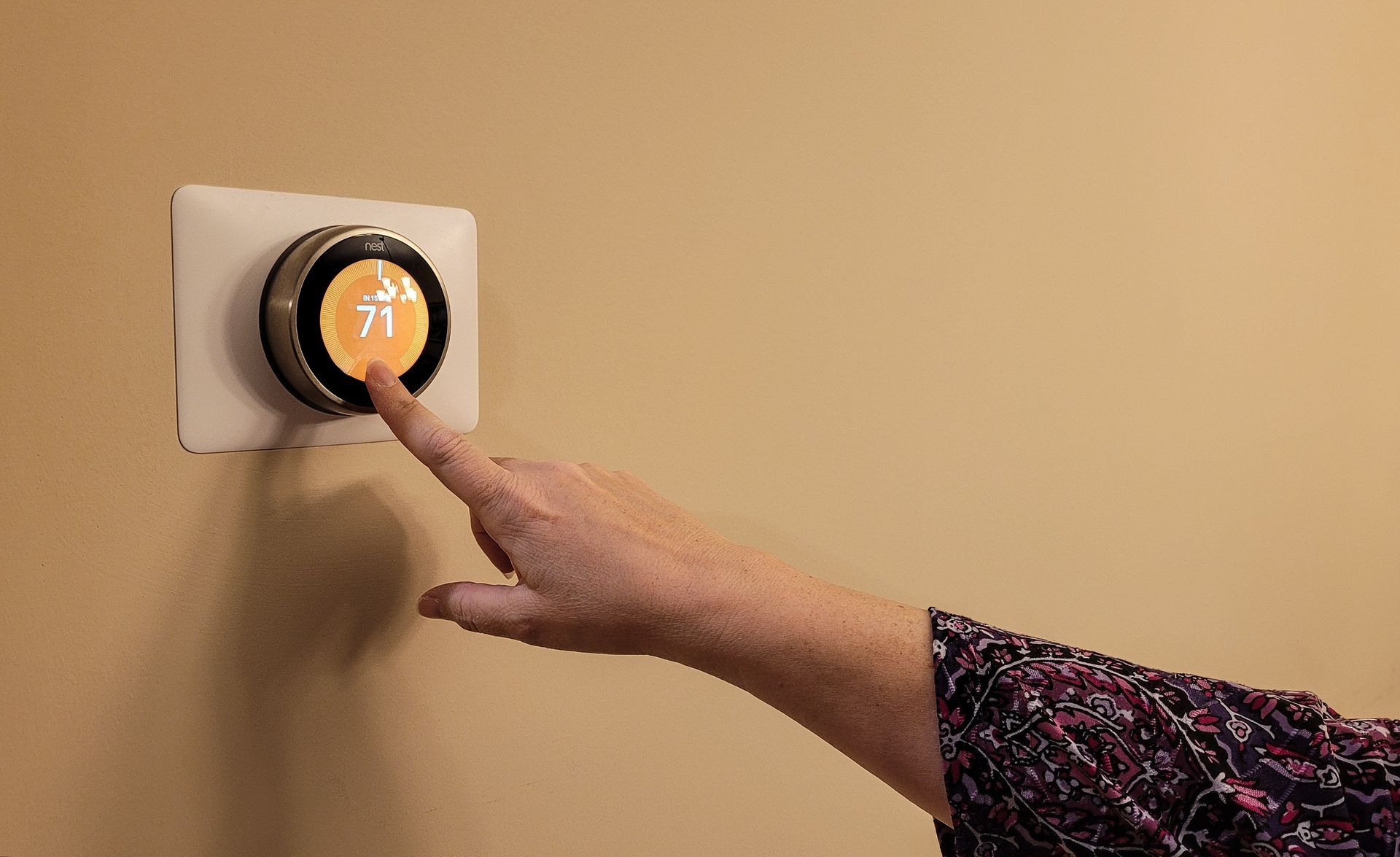 A hand adjusting a smart thermostat on a beige wall, with the temperature displayed as 71 degrees.