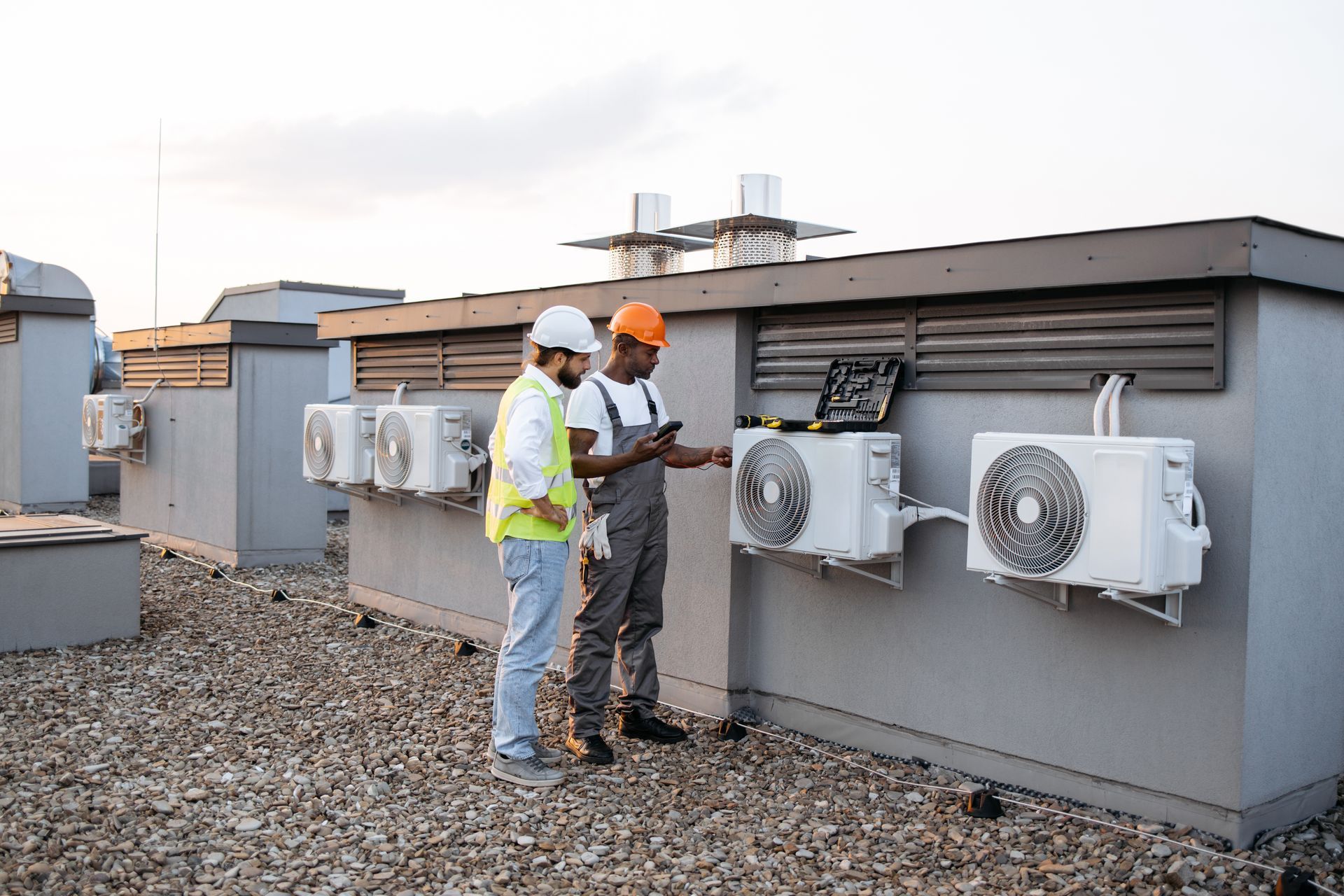 Two HVAC technicians on a rooftop examine air conditioning units. One uses a phone, the other wears a hard hat.