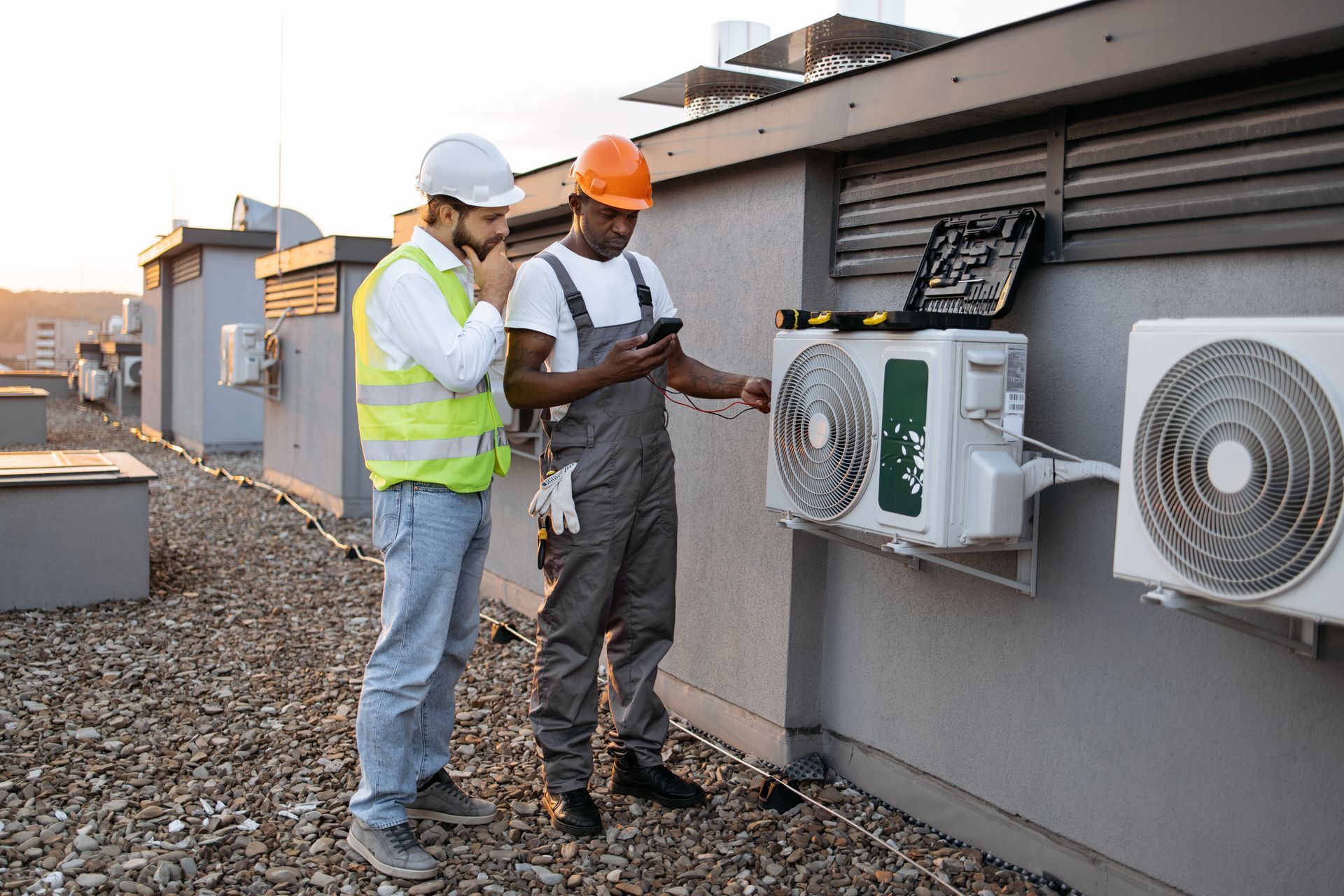Two HVAC technicians on a rooftop, examining an air conditioning unit. One looks at a phone, the other looks thoughtful.