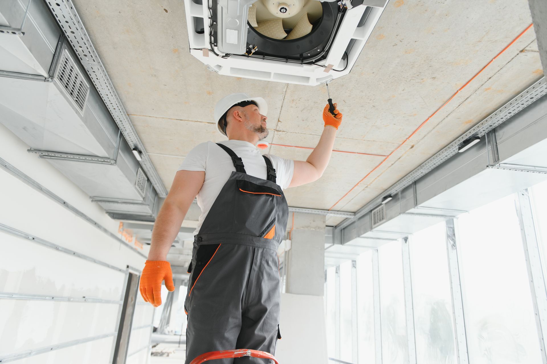 Man in overalls and hardhat repairs ceiling AC unit with a screwdriver, indoors.
