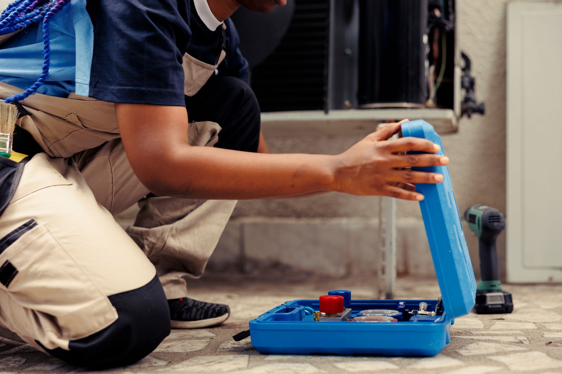 HVAC technician kneeling, opening a blue toolbox outdoors near AC unit.